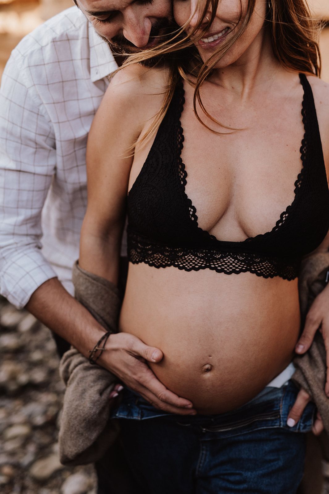 Pregnant woman in black lace bralette and jeans cradling baby bump with partner outdoors.