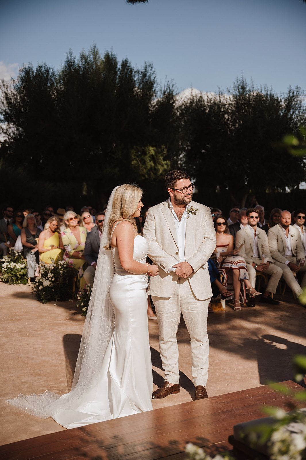 A bride and groom hold hands at an outdoor wedding ceremony surrounded by guests and lush green trees.