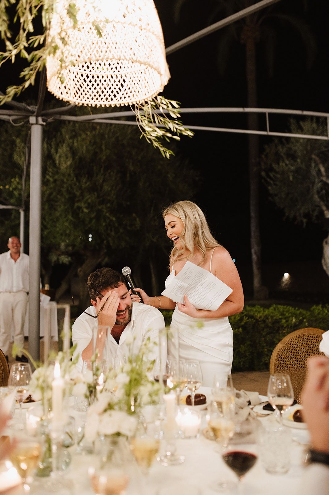 Blonde bride in white dress giving speech with microphone at elegant outdoor wedding reception.