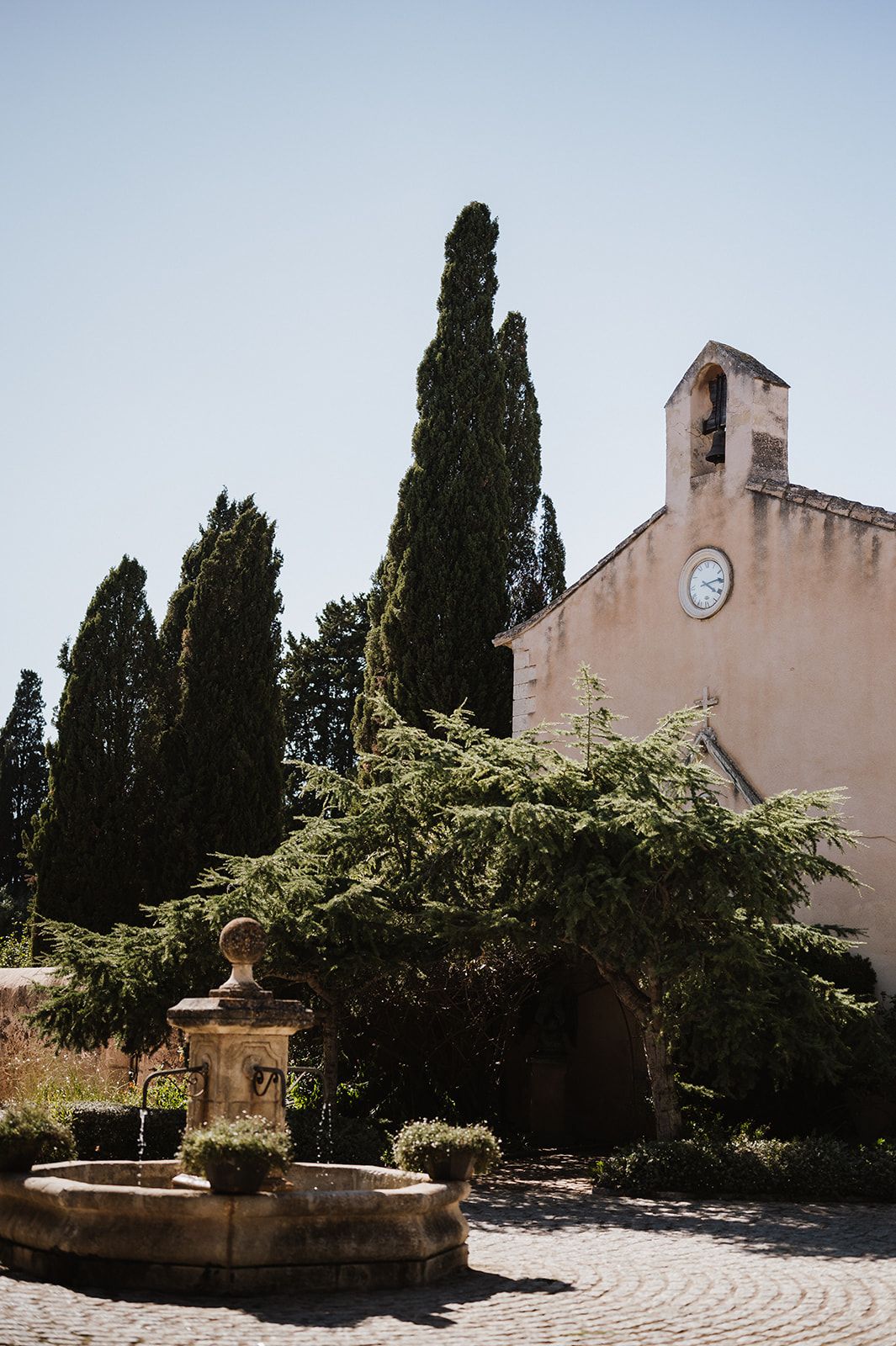 Rustic stone chapel with a clock tower, cypress trees, and a classic fountain on cobblestone grounds.