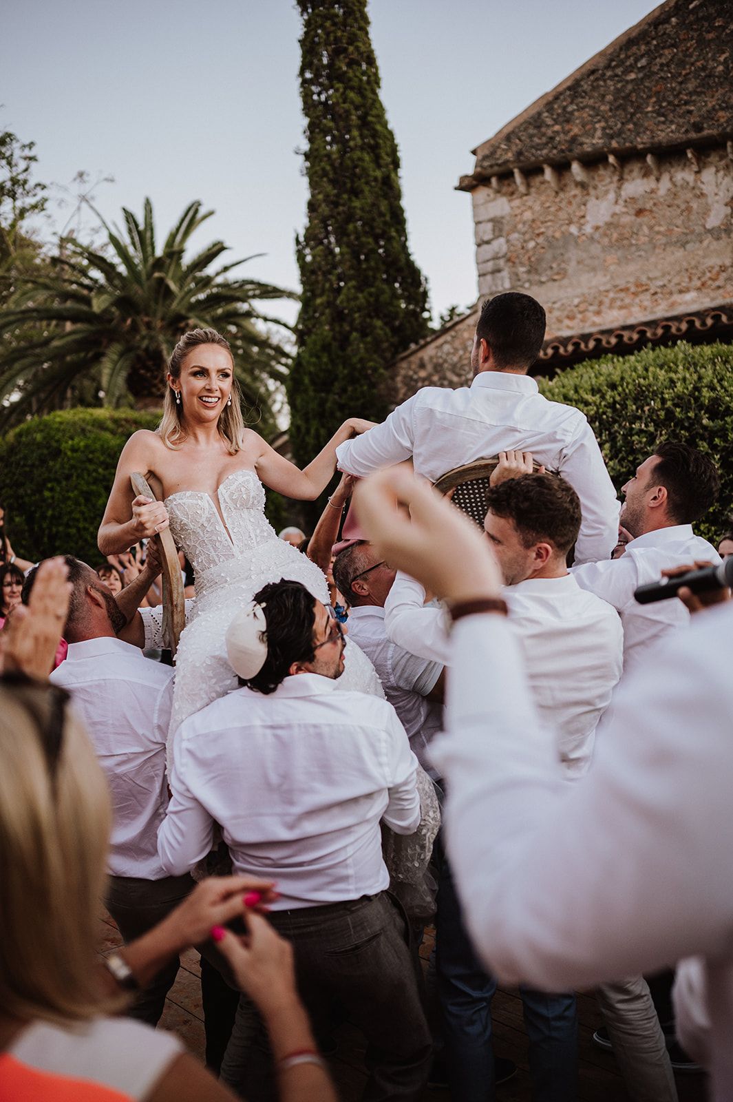 Bride lifted by groomsmen in white shirts during lively outdoor wedding celebration.