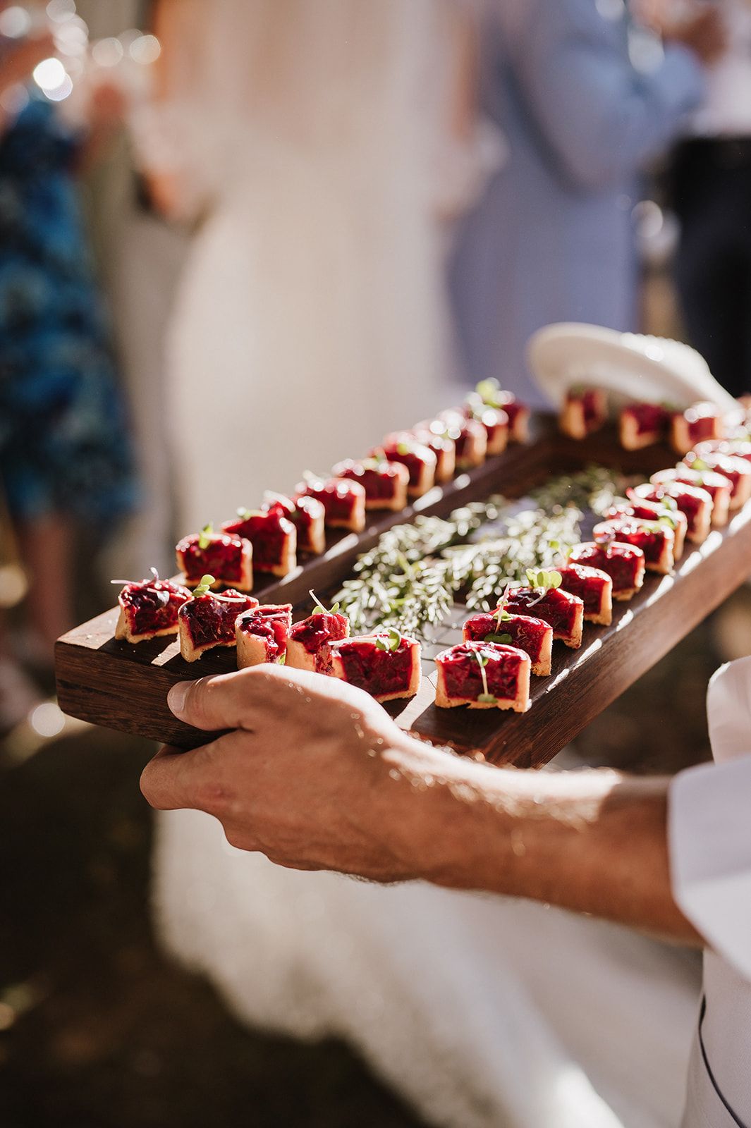 Server holds wooden tray of elegant red berry tartlets at outdoor wedding reception.