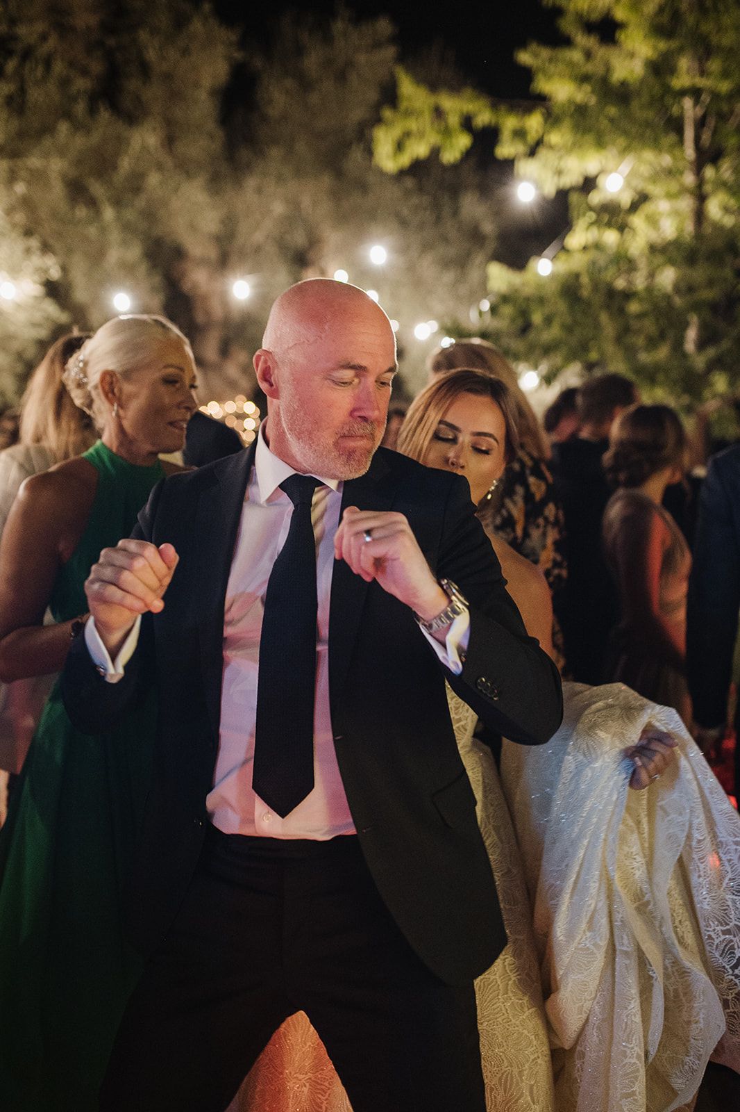 Bald man in suit dancing with bride at lively outdoor wedding reception under string lights.