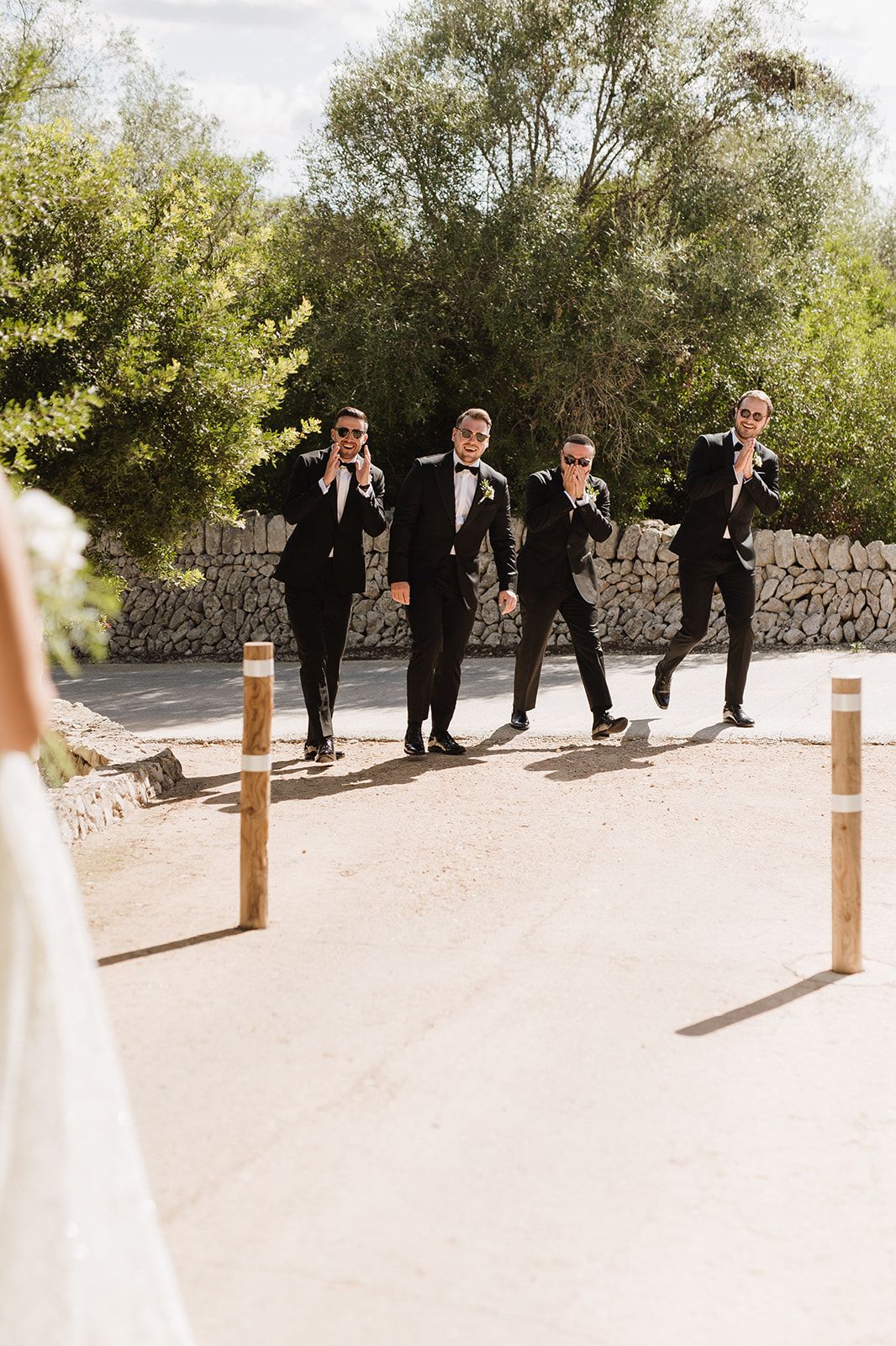 Four groomsmen in black suits react with joy upon seeing the bride at an outdoor wedding ceremony.