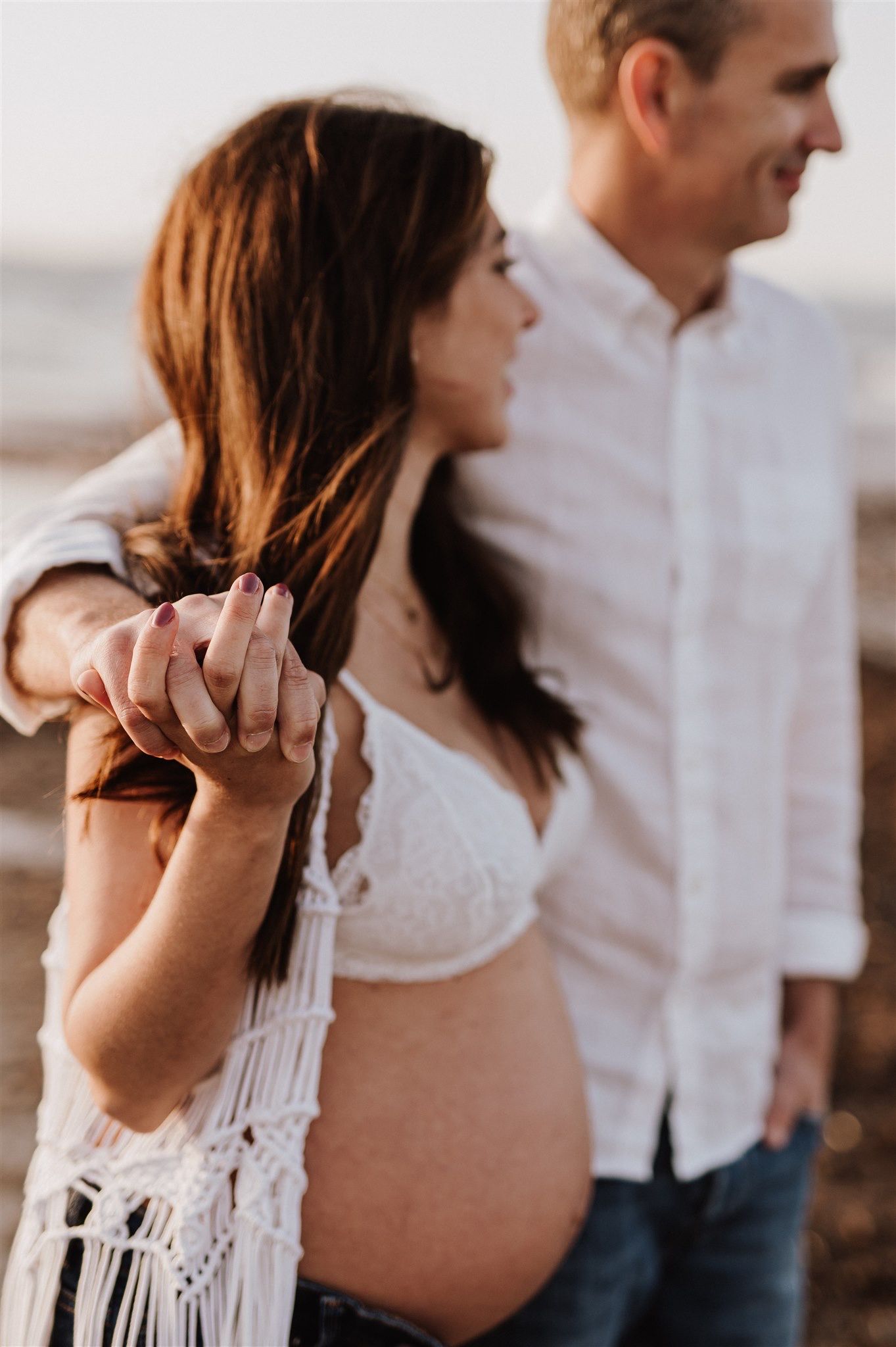 Pregnant woman in white lace bralette holds hands with man in white shirt during golden hour maternity photo session.