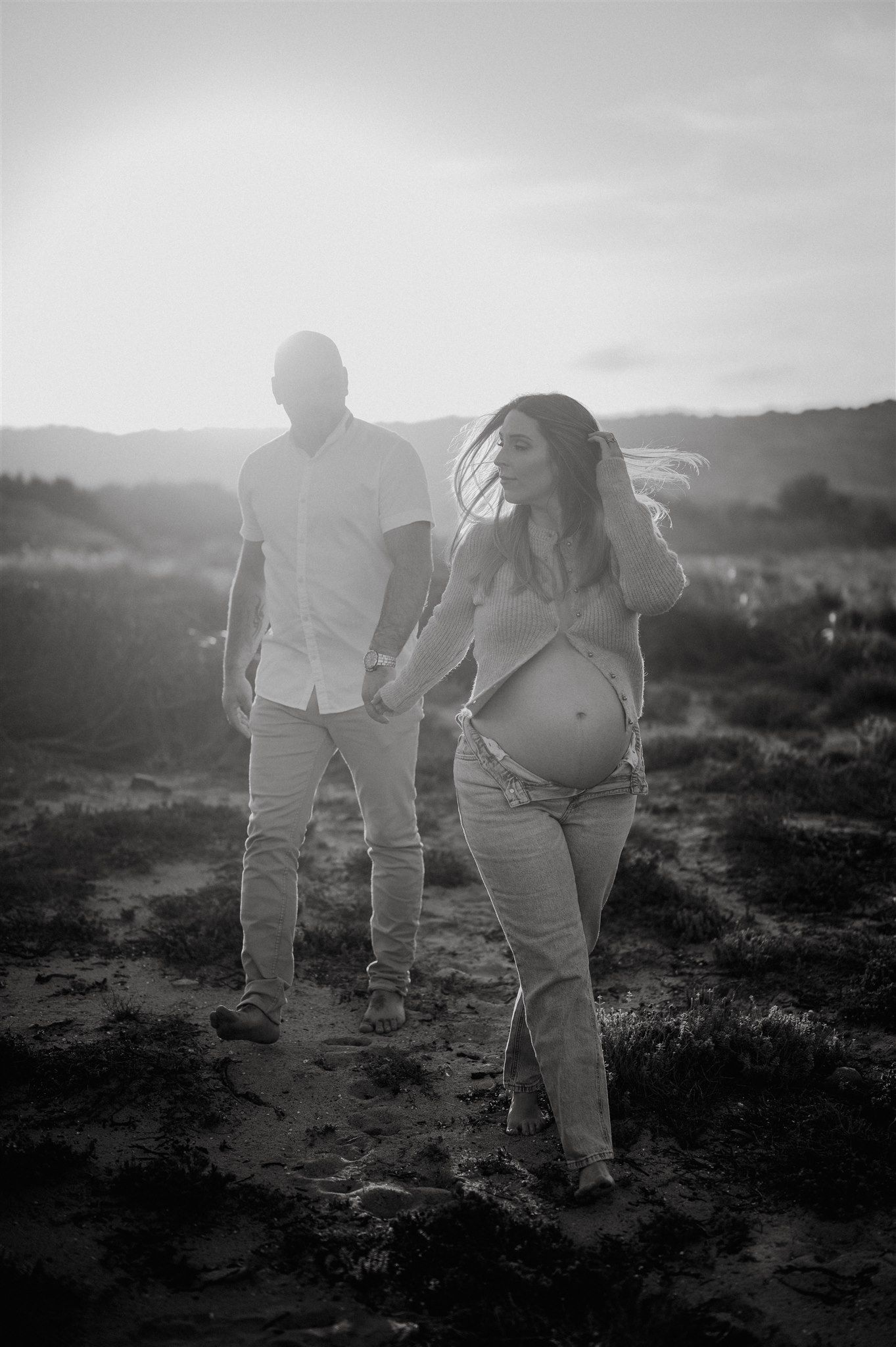 Black and white photo of expectant couple holding hands at sunset during outdoor maternity photoshoot on sandy terrain.
