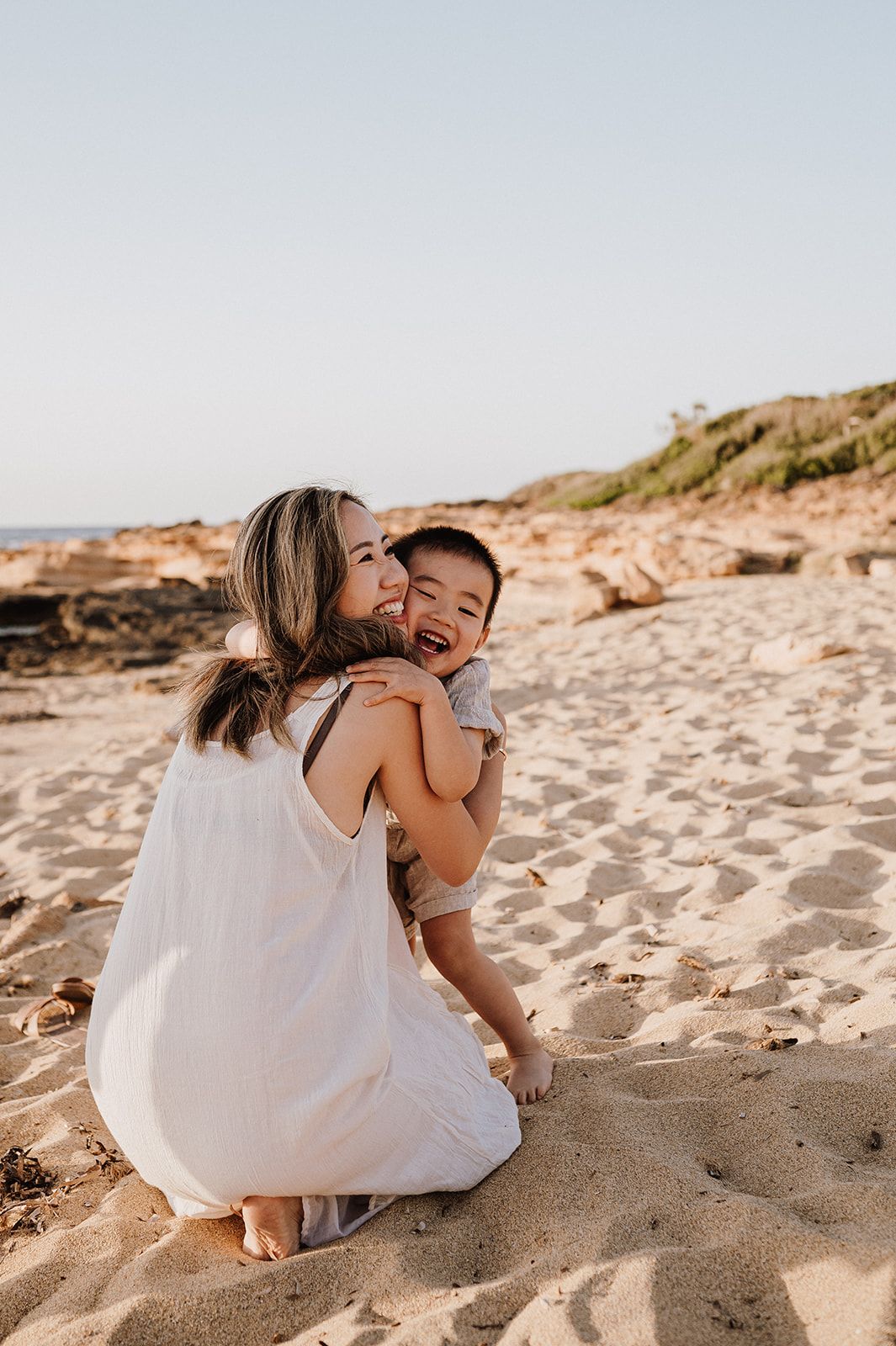 A mother in a white dress hugs and kisses her laughing toddler on a sandy beach at golden hour.