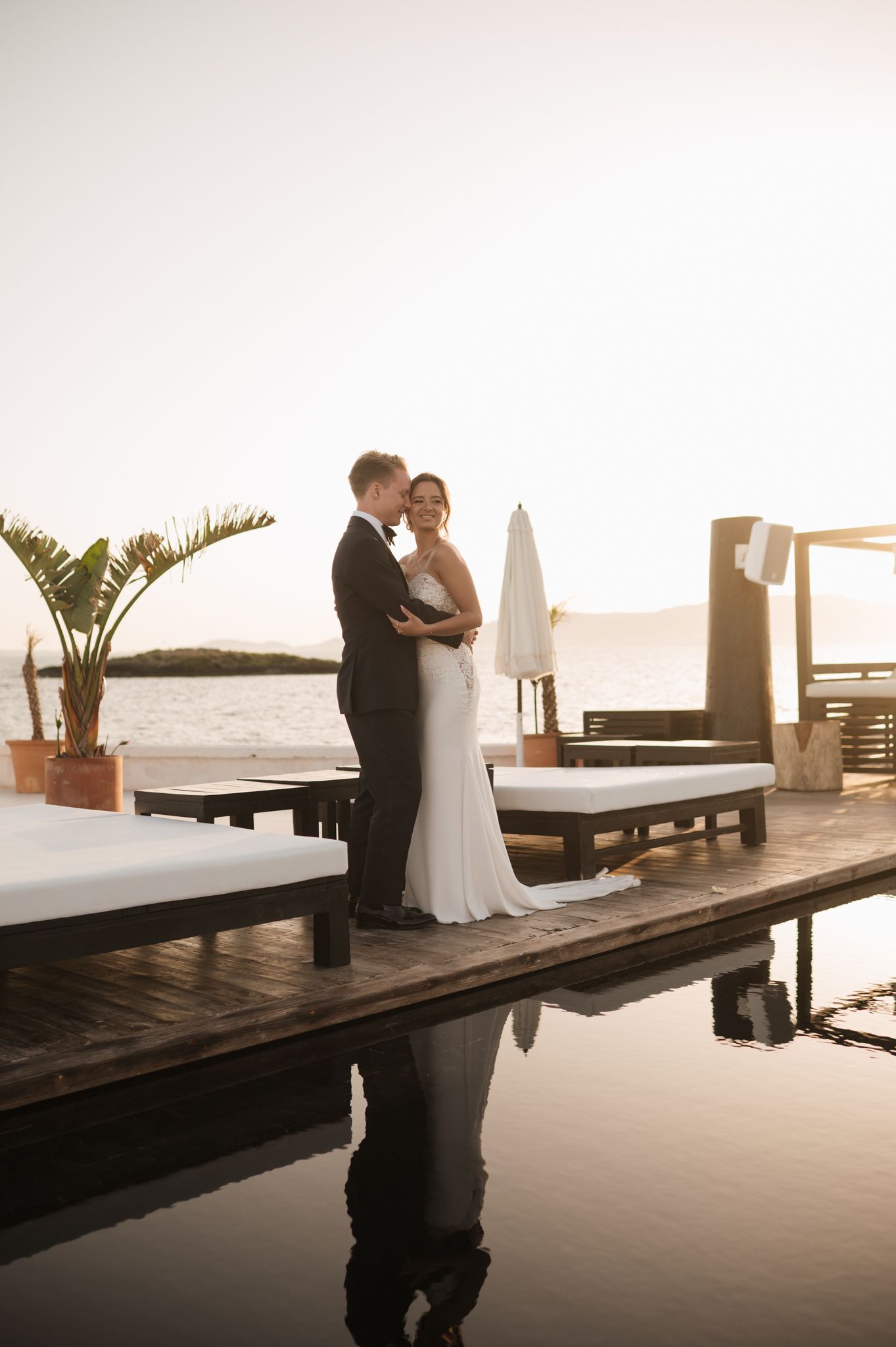 Newlyweds share romantic kiss on wooden dock at sunset, reflected in calm poolside water below.
