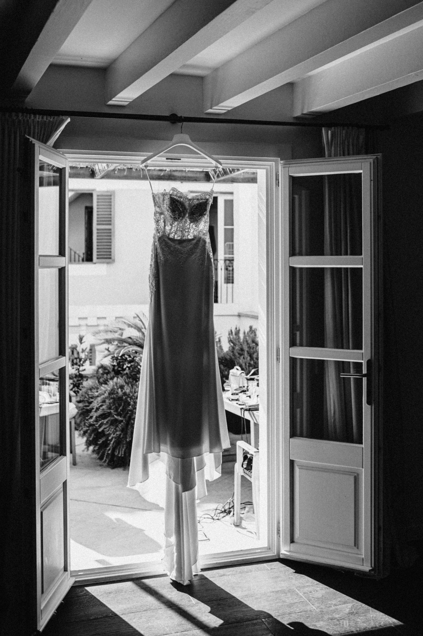 Elegant wedding dress hanging on a hanger in an open doorway with a sunlit courtyard visible beyond.