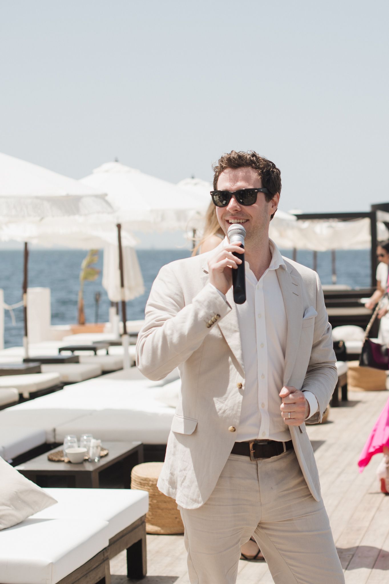 Stylish man in beige suit speaking into microphone at an outdoor seaside wedding event.