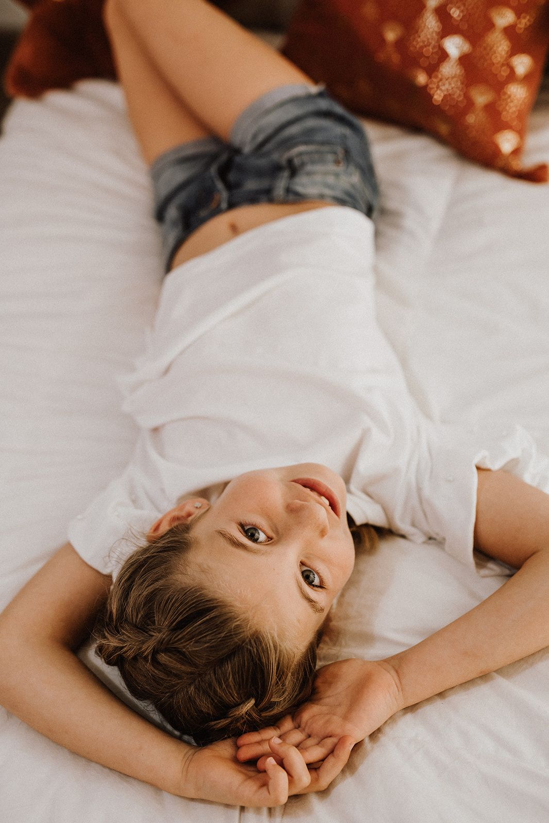 Young girl lying on white bed wearing denim shorts and white shirt, looking up at camera with a smile.