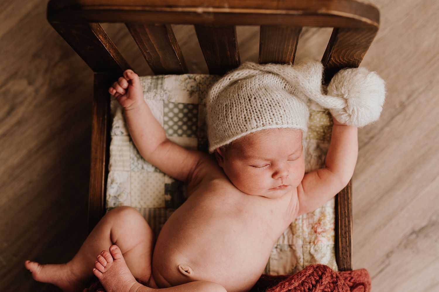 Sleeping newborn baby wearing a knit pom-pom hat lying in a small wooden bed.