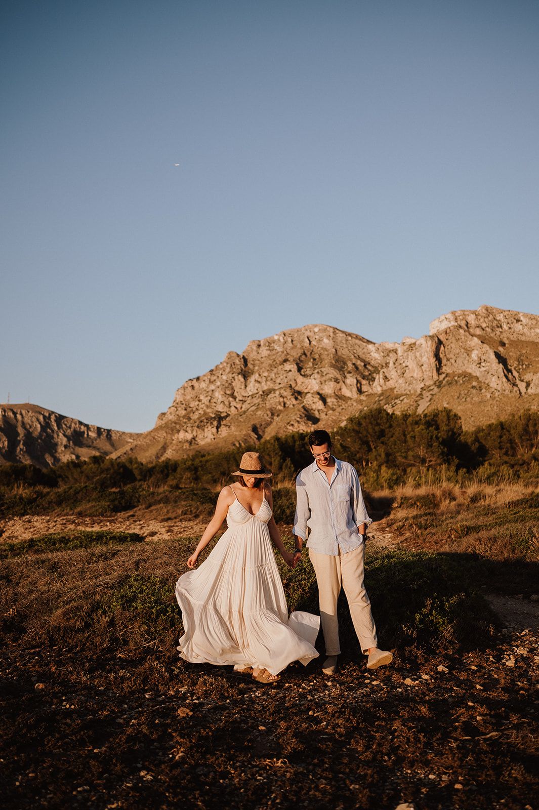 Couple walking in golden hour sunlight with mountain backdrop, woman in flowing white dress and straw hat, man in light outfit.