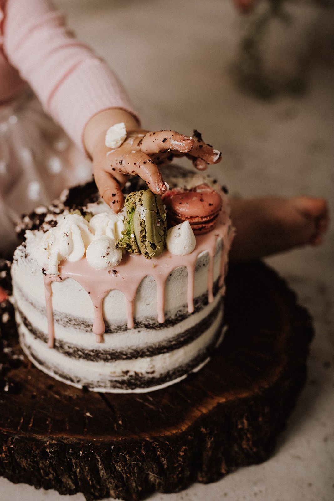Child's hand reaching into a decorated naked cake with pink drip frosting and macarons on a rustic wood stand.
