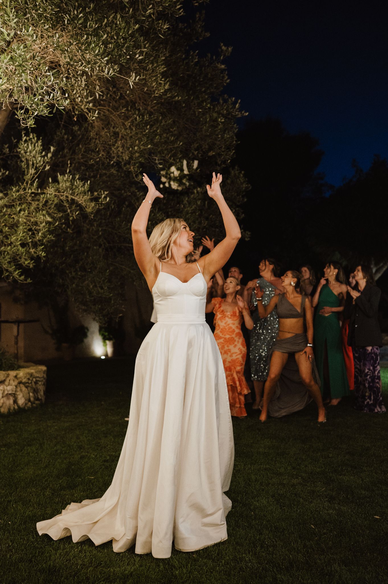 Bride in white gown tosses bouquet with arms raised at an outdoor evening wedding reception celebration.