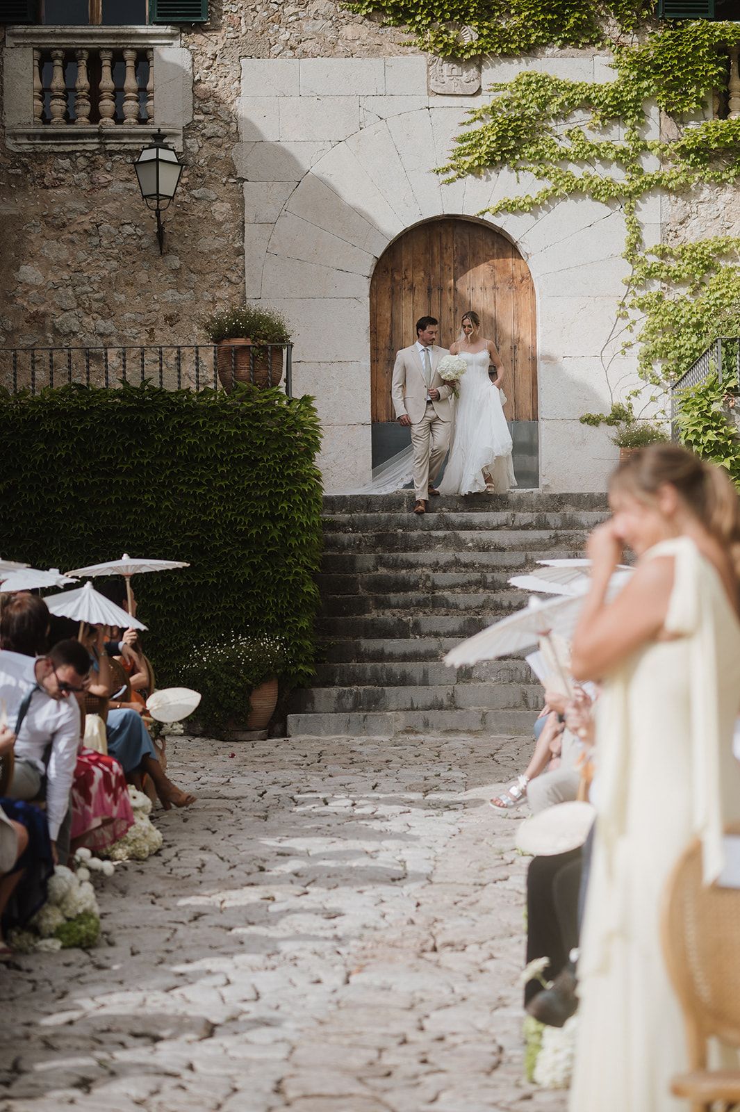 Bride and groom descend stone steps at rustic villa wedding ceremony while guests watch from cobblestone aisle.