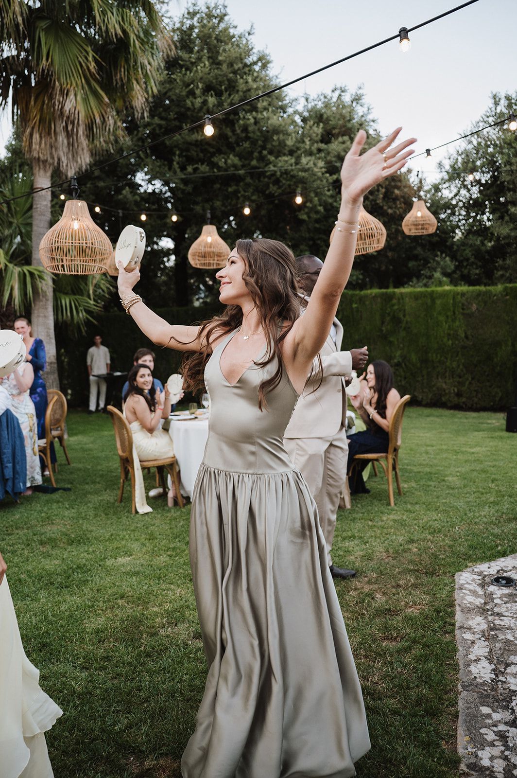 Joyful woman in sage satin gown dances with arms raised at elegant outdoor garden wedding reception.