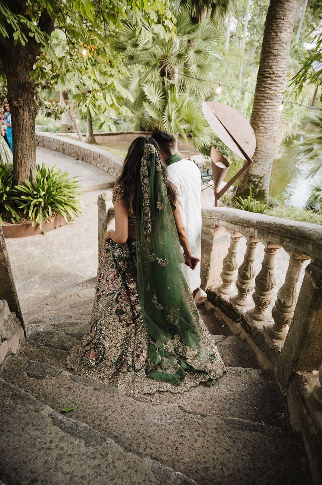 Bride in embroidered green lehenga with matching dupatta walking up stone stairs at outdoor venue.