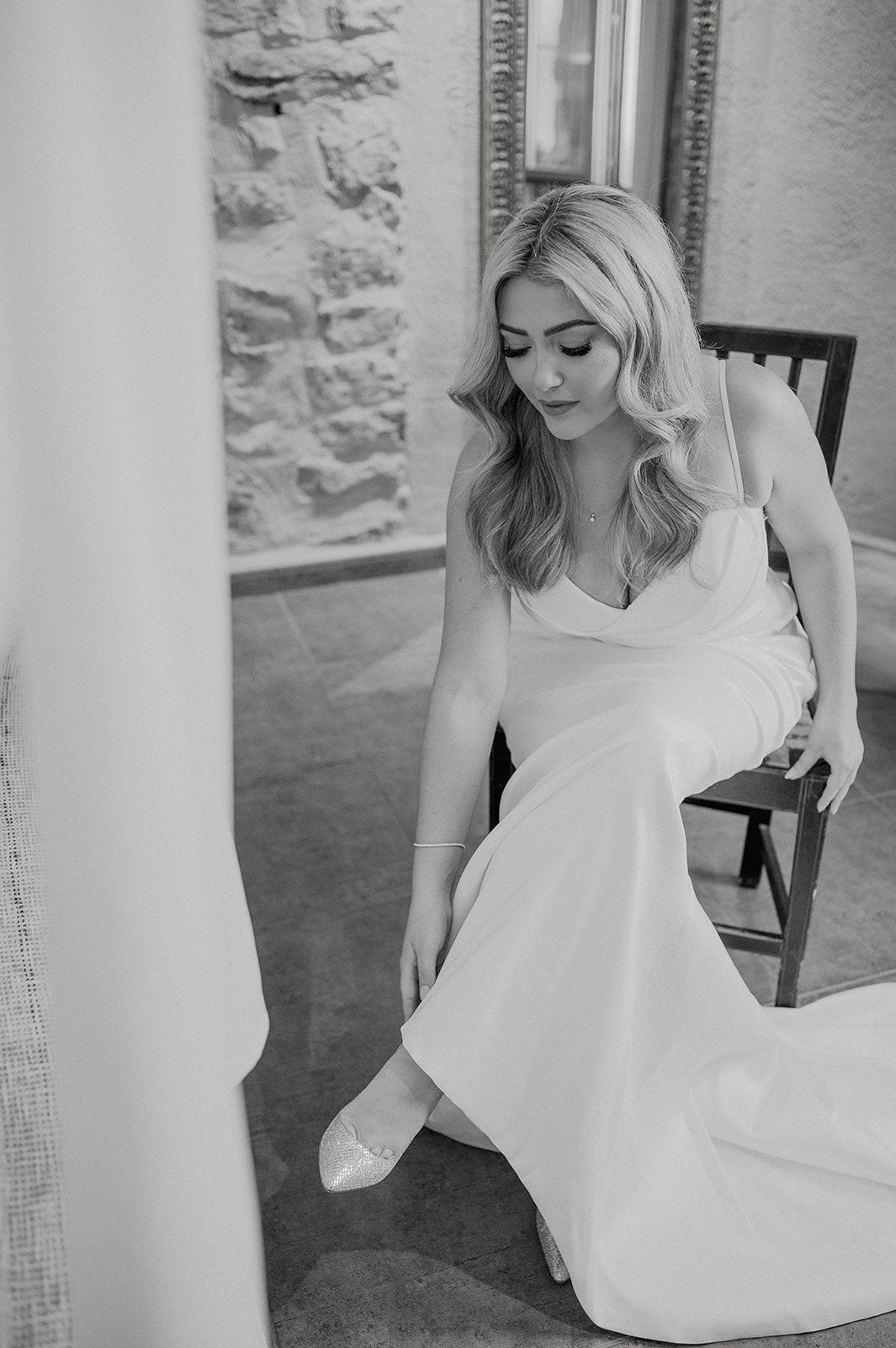 Elegant bride in a sleek white wedding gown putting on her shoes in a black and white photo.