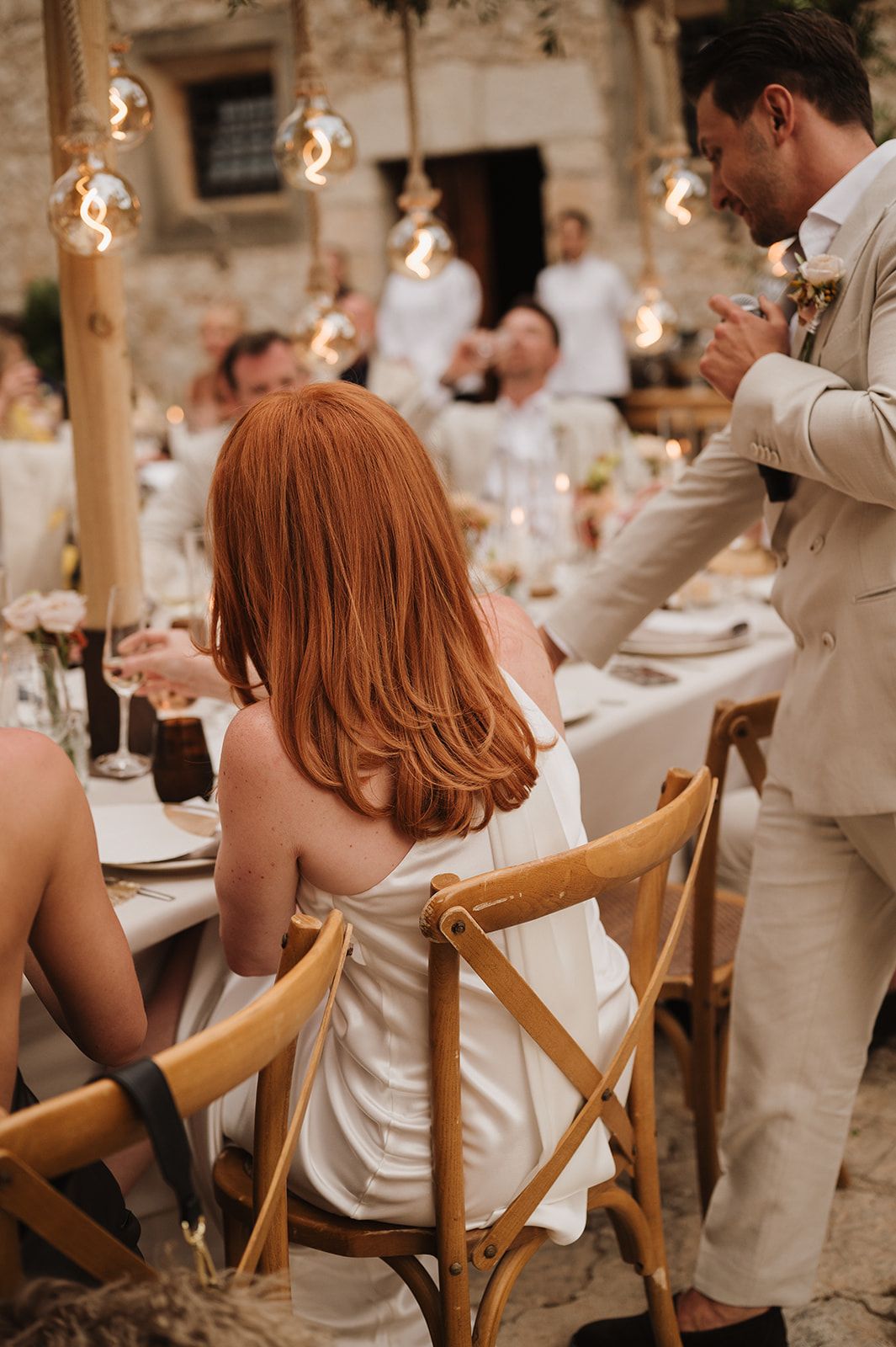 Red-haired bride in white satin dress seated at outdoor wedding reception with Edison bulb lighting.