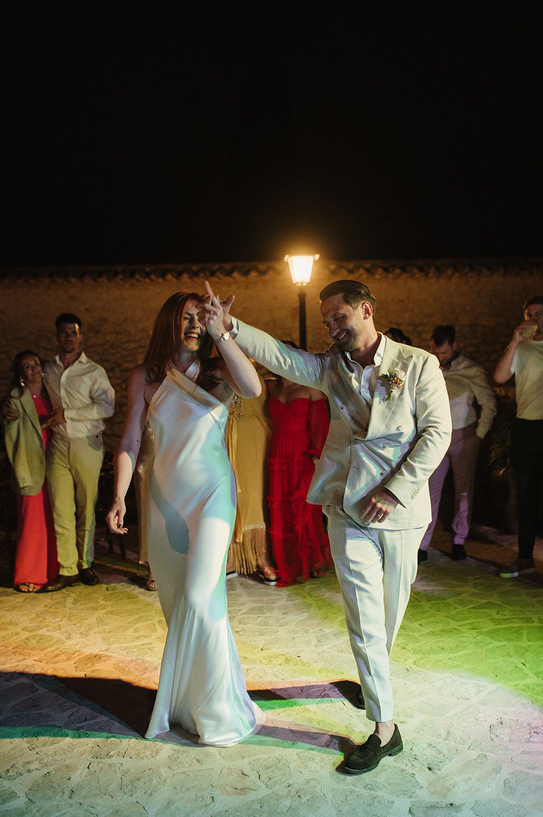 Bride in white satin gown and groom in beige suit sharing first dance under torch light at night.