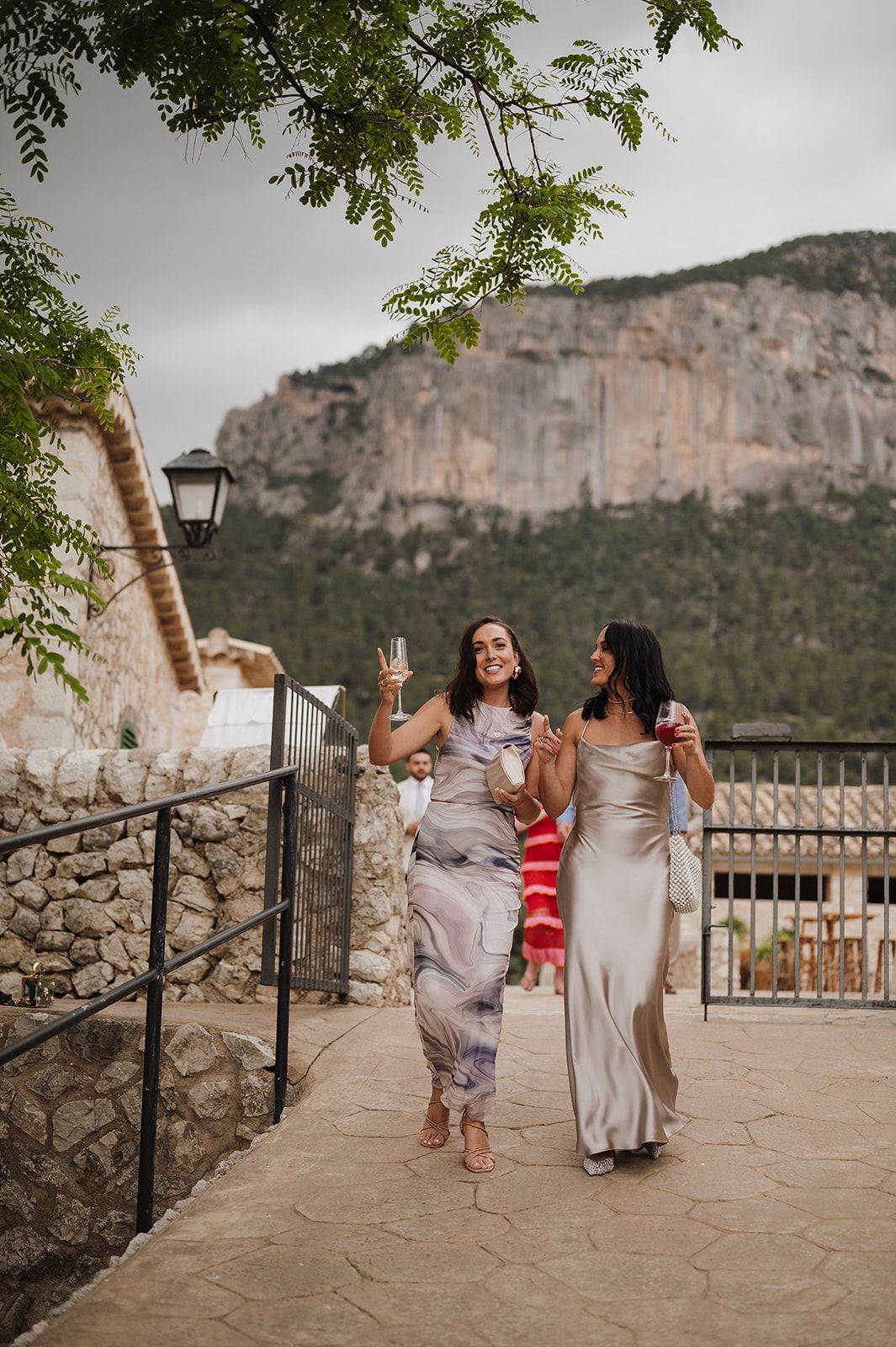 Two elegantly dressed women walking along a stone terrace with a dramatic rocky mountain backdrop.