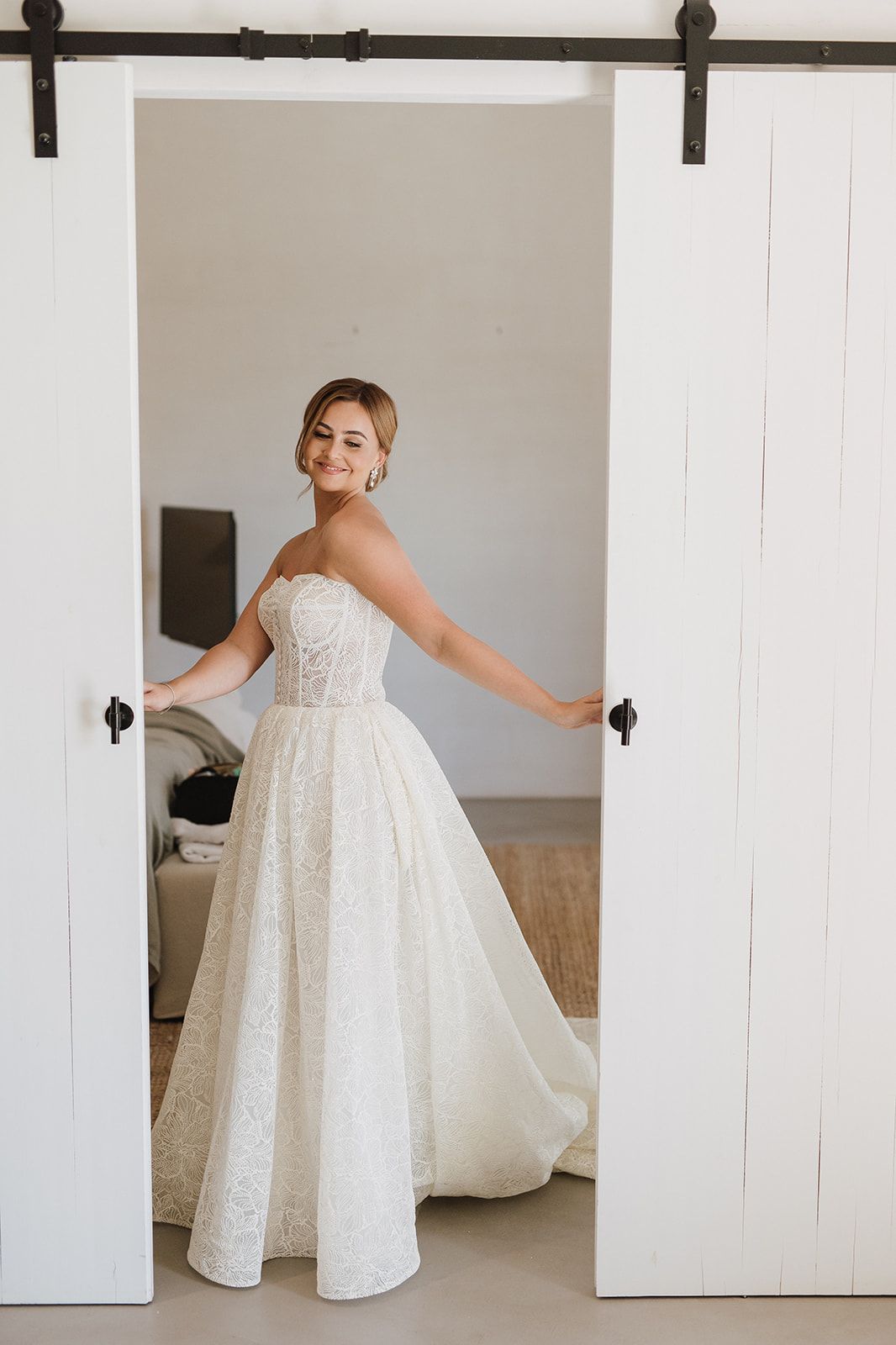 Elegant bride in strapless lace ball gown smiles while standing in barn-style doorway.