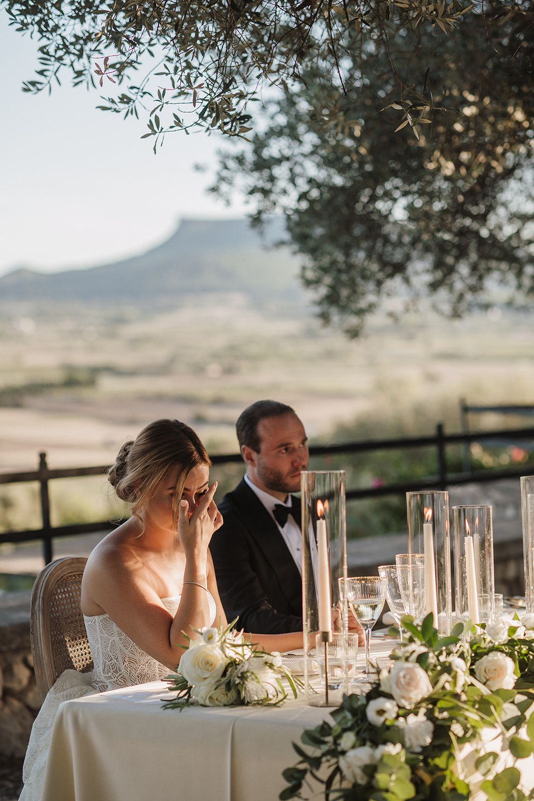 Bride and groom sit at elegant outdoor sweetheart table with white floral arrangements and candles, scenic Tuscan landscape behind.