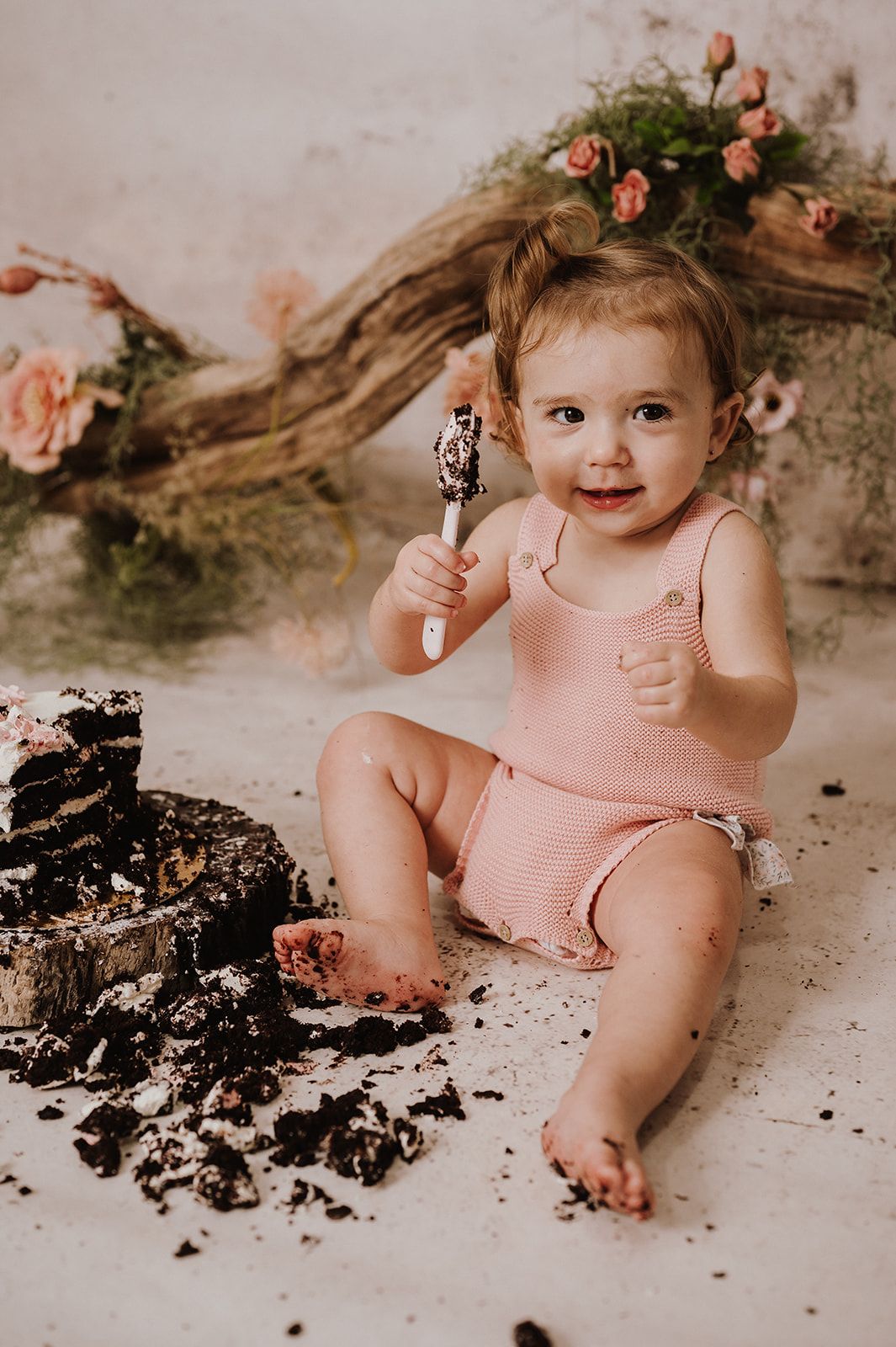 Baby girl in pink romper smashes first birthday cake, holding fork and smiling during cake smash photo session.