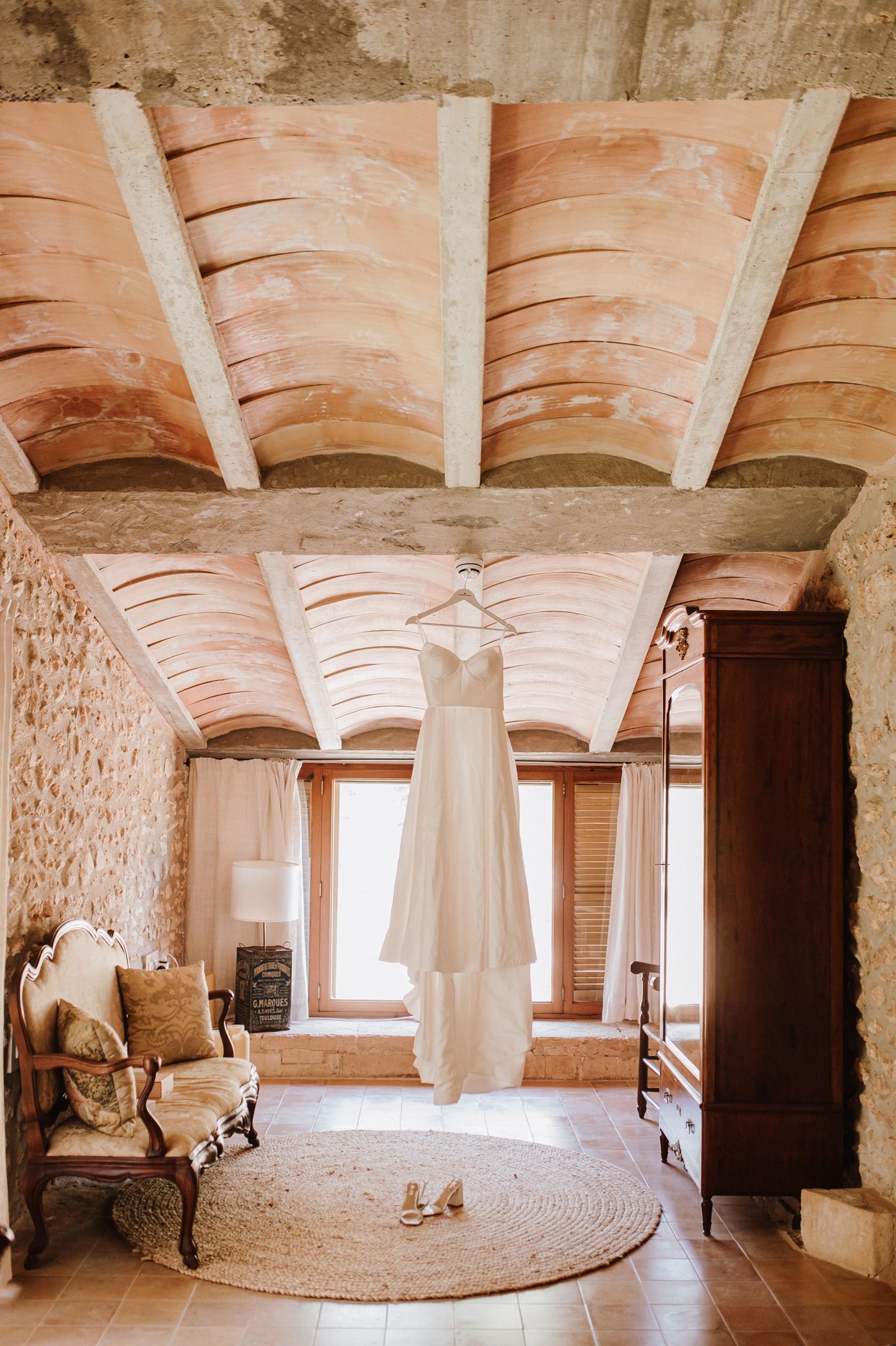 White wedding dress hanging in a rustic stone room with arched ceiling beams, wooden furniture, and a round jute rug.