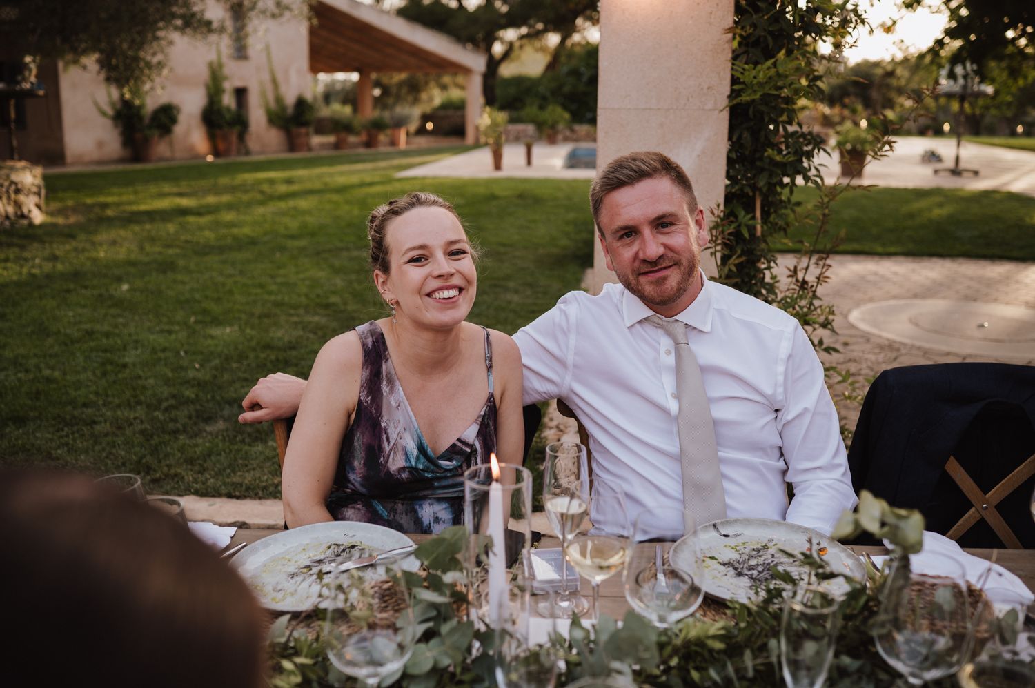 Smiling couple enjoys an outdoor wedding reception dinner seated at a candlelit table in a garden.
