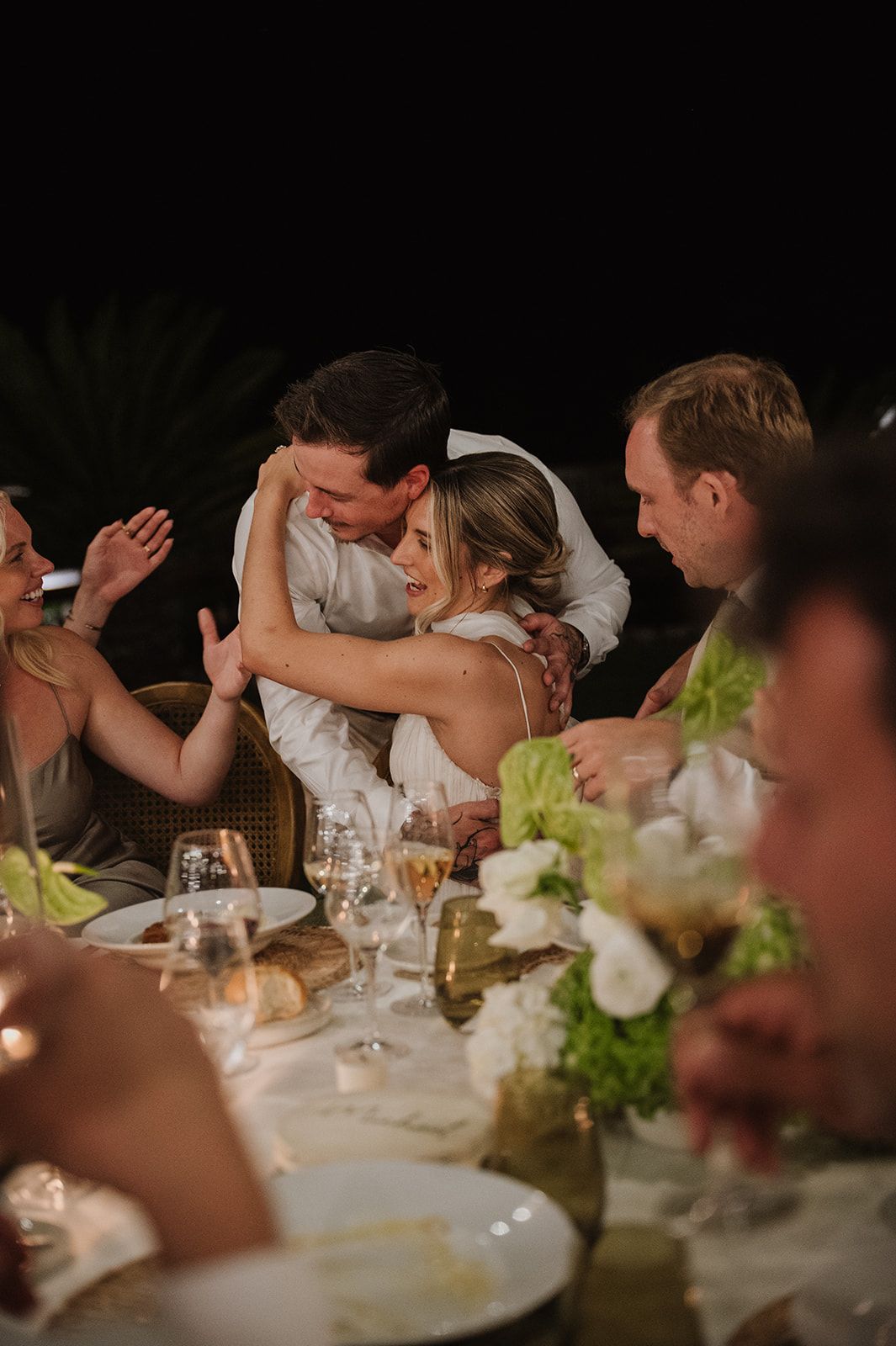 Bride and groom sharing tender embrace at candlelit wedding reception dinner table with guests.