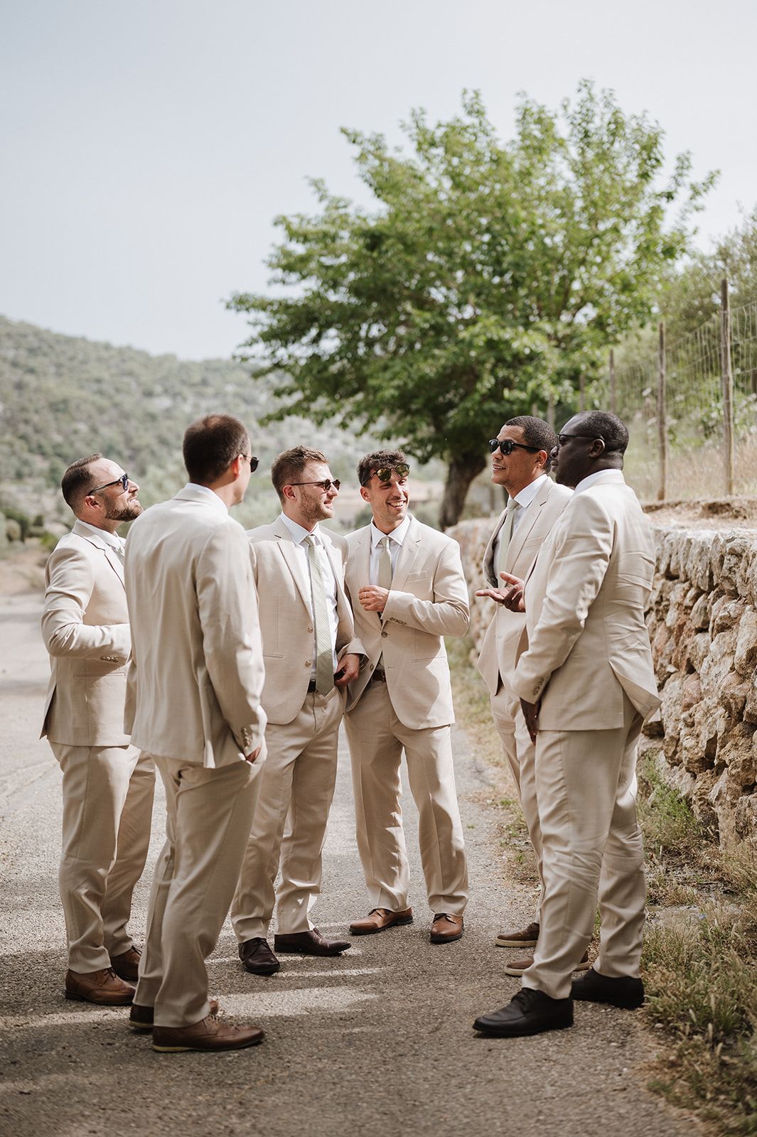 Six groomsmen in matching tan suits socializing outdoors near a stone wall with green hills in the background.