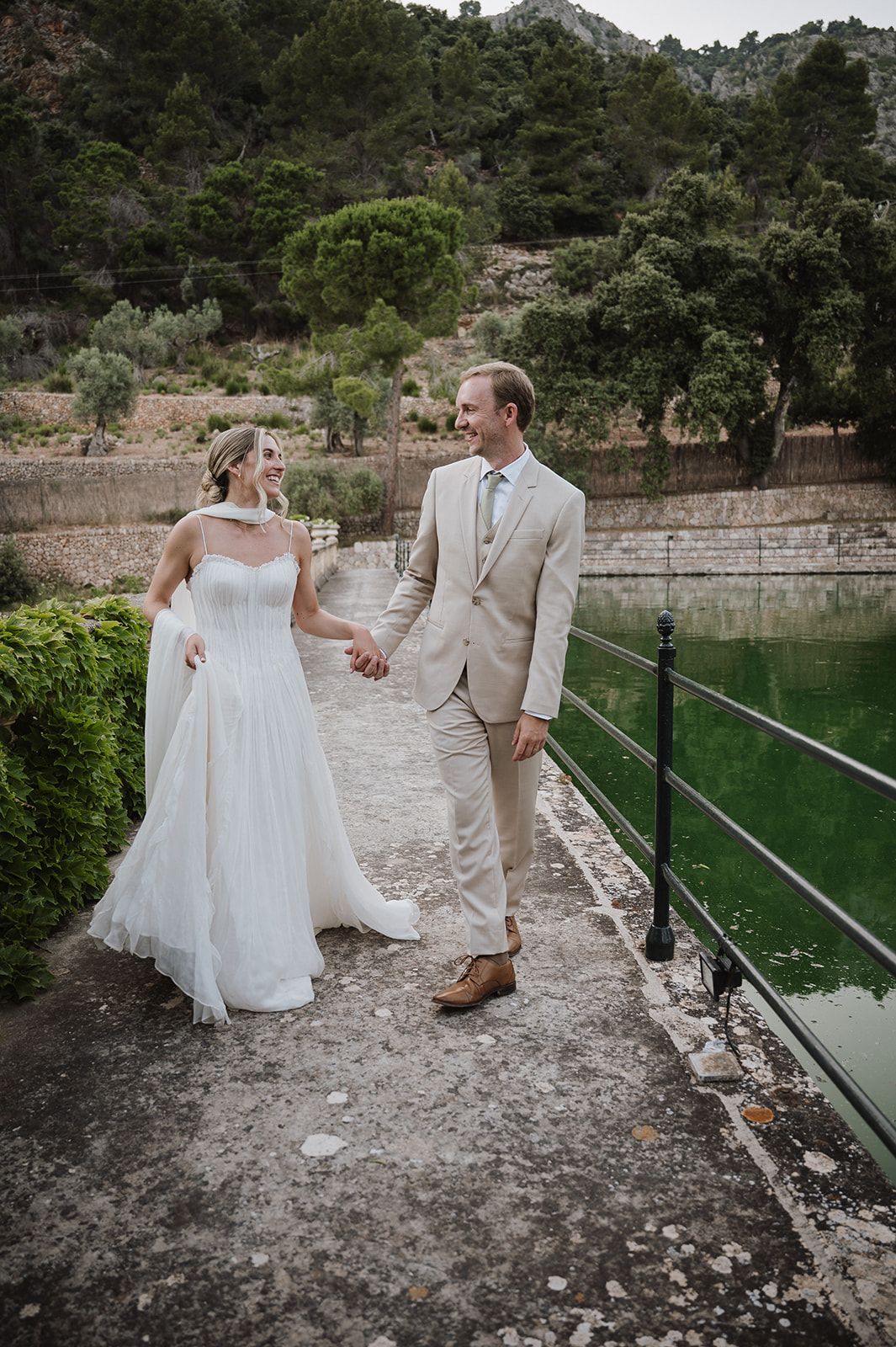 Bride in white tulle gown and groom in beige suit hold hands walking along a scenic waterside path.