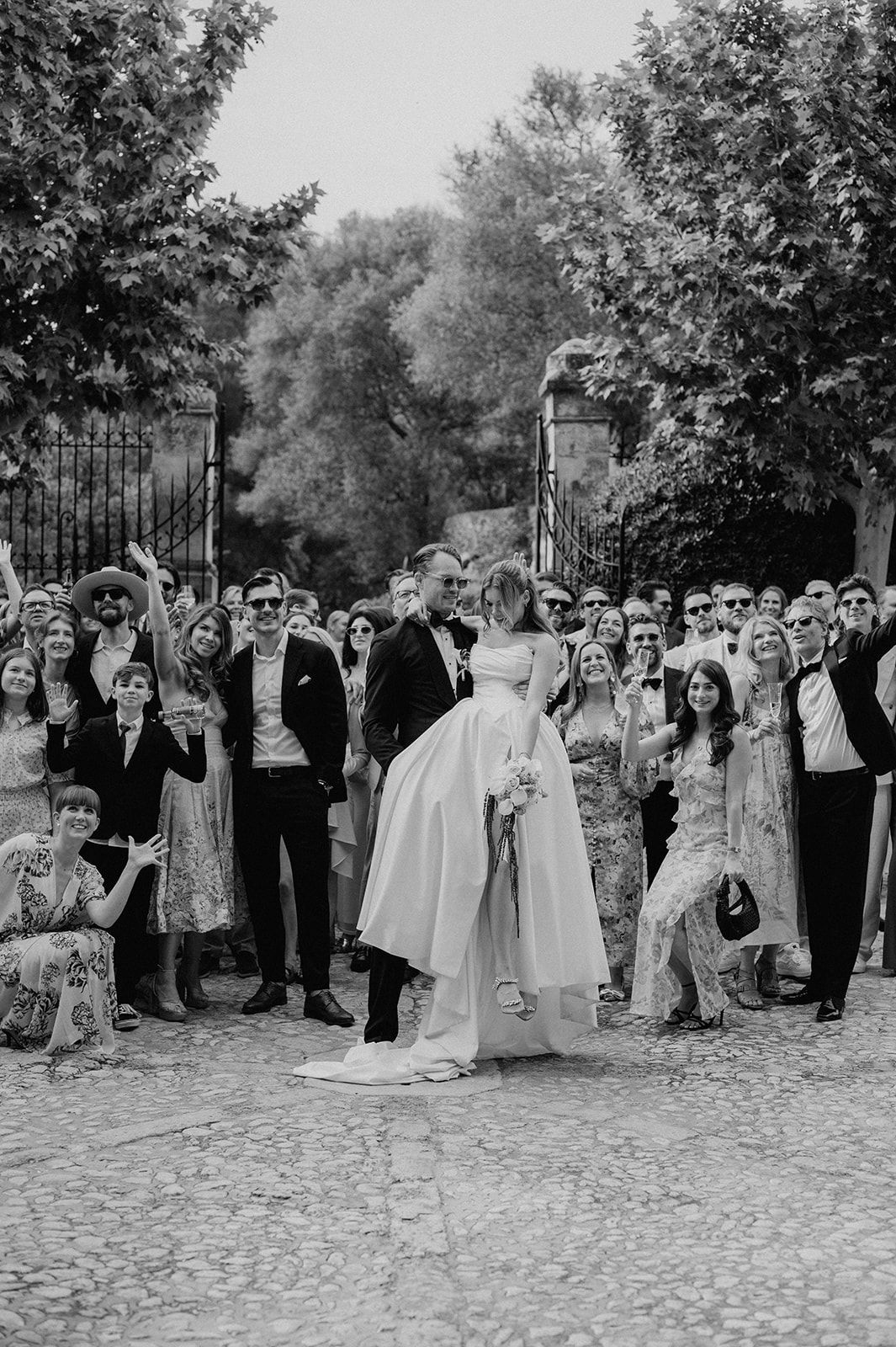 Black and white wedding photo of couple kissing surrounded by cheering guests outdoors near iron gates and trees.