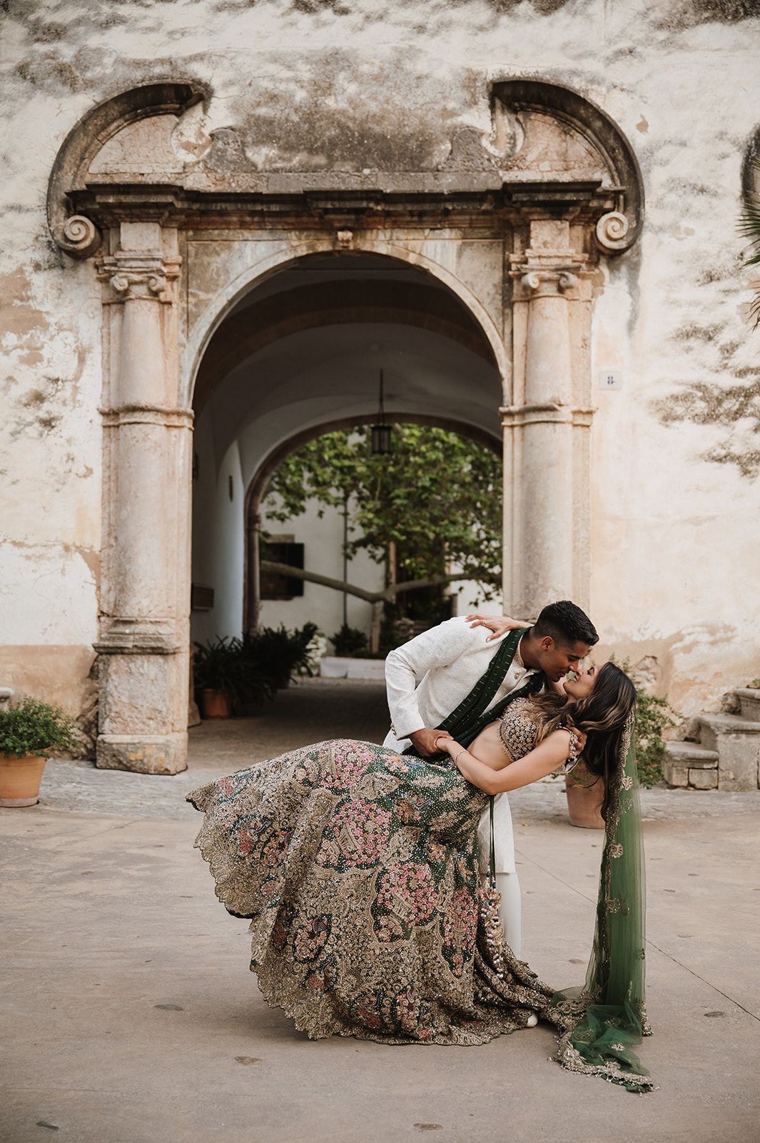 Couple in Indian wedding attire share a romantic dip kiss before a stone archway.