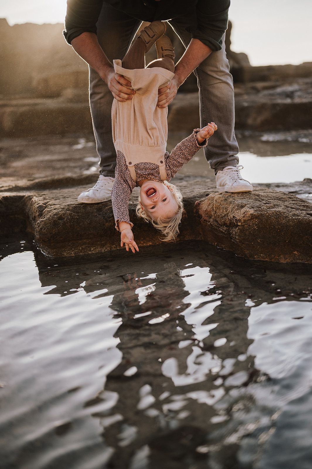 Adult playfully holds laughing toddler upside down over a reflective tidal pool on rocky coastal shore at golden hour.