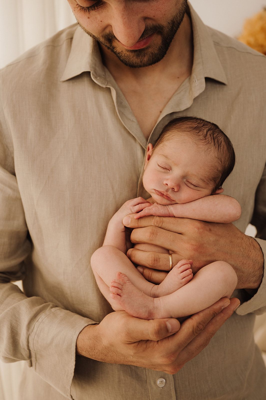 Father in linen shirt gently cradles sleeping newborn baby in his hands, looking down lovingly.