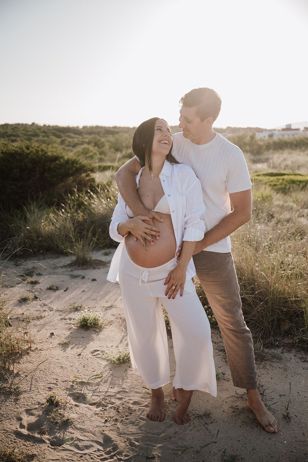 Couple embracing on sandy beach dunes at sunset, pregnant woman in white outfit smiling joyfully.