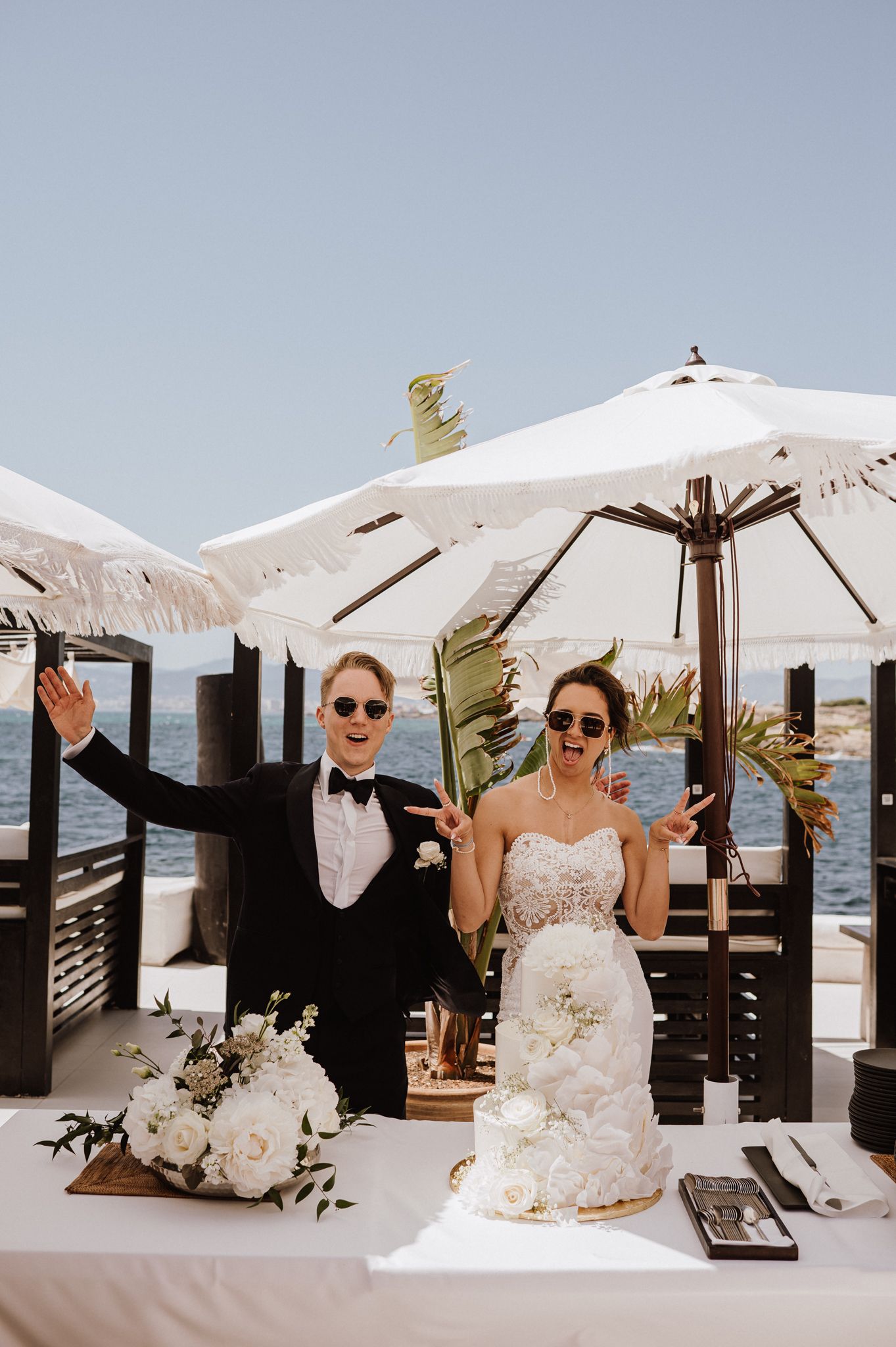 Joyful bride and groom celebrating at wedding cake table by the sea under white umbrellas.