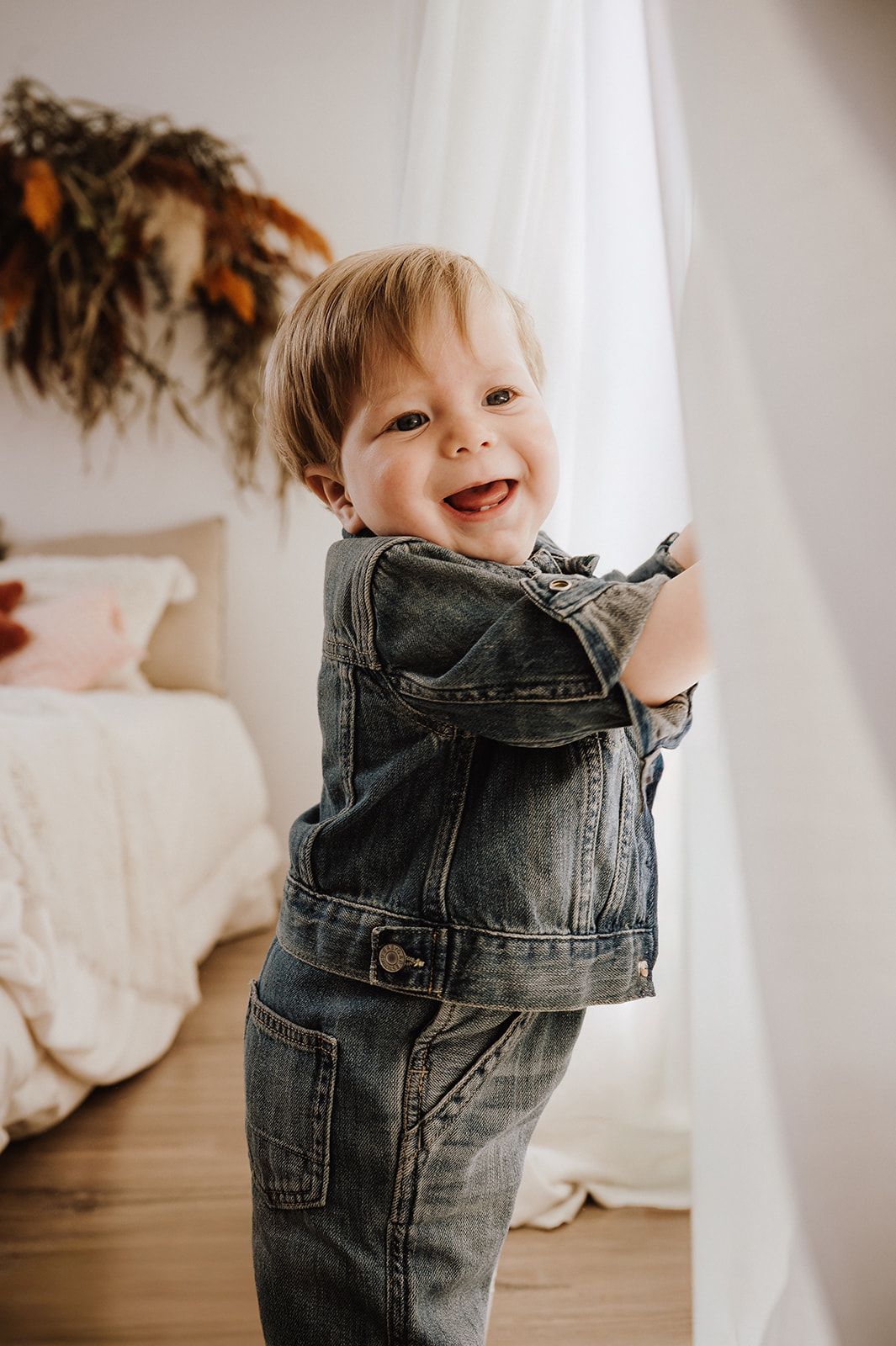 Happy toddler boy in denim jacket and jeans smiling while playing with white curtain in bedroom.