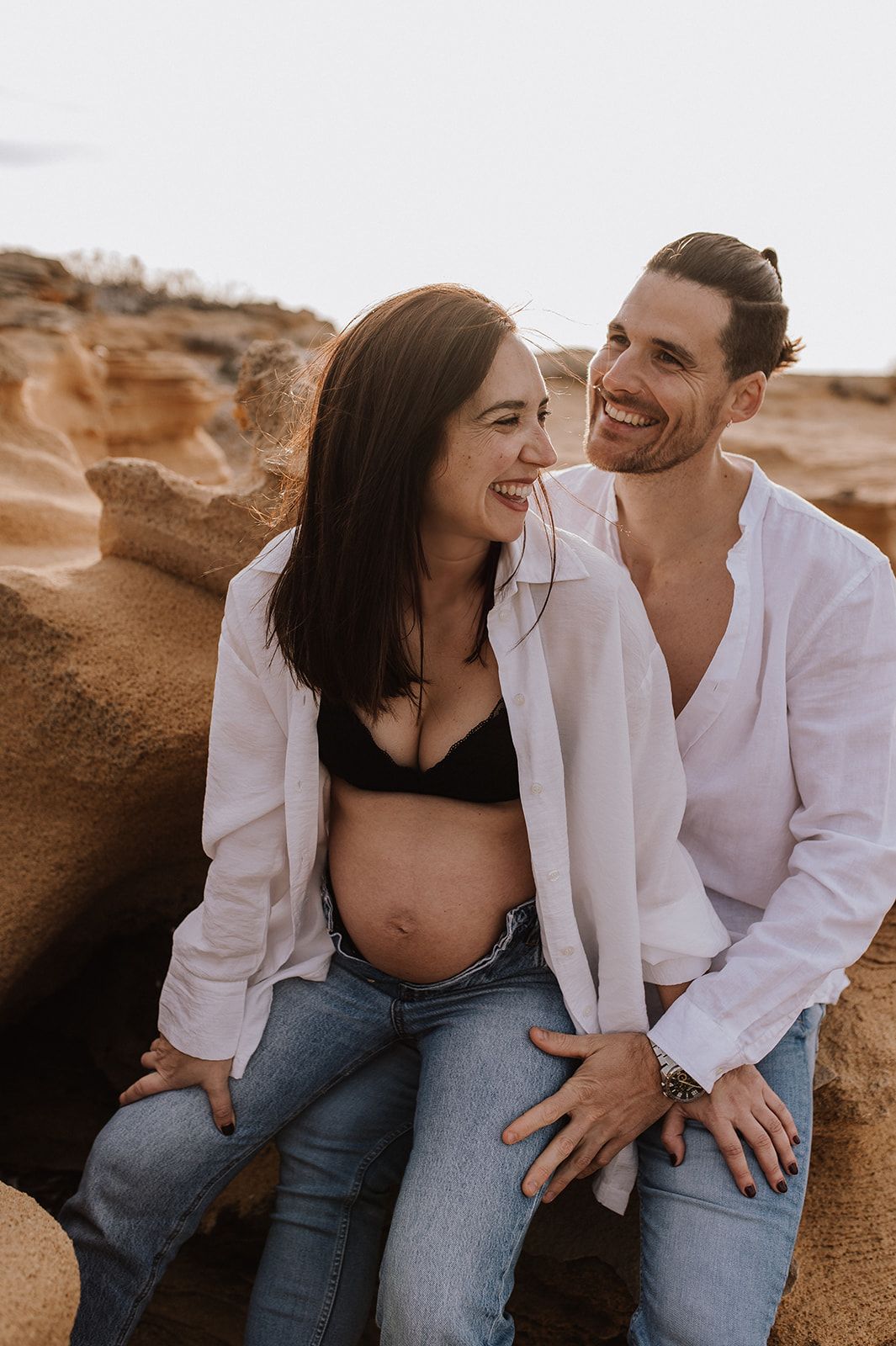 Happy couple in white shirts, pregnant woman showing baby bump, sitting on rocks outdoors, laughing together.