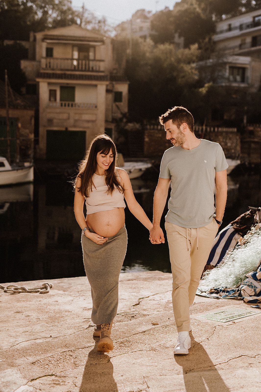 Pregnant couple holding hands and walking along a sunny Mediterranean harbor, smiling at each other during maternity photoshoot.