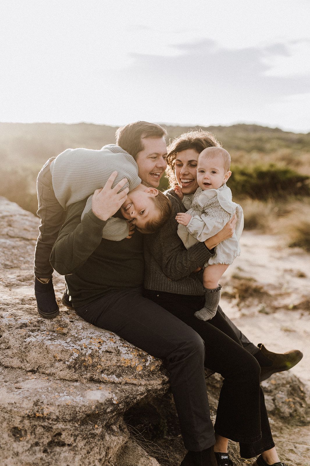 A happy family of four sits on a rock outdoors, parents cuddling two young children in warm golden sunlight.