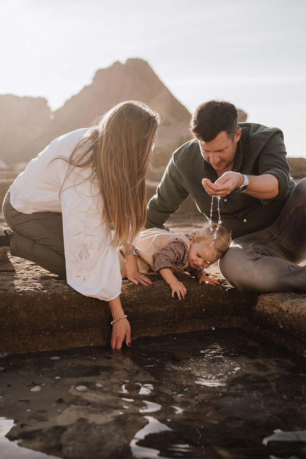 Family with toddler playing with water in a coastal tide pool at golden hour.