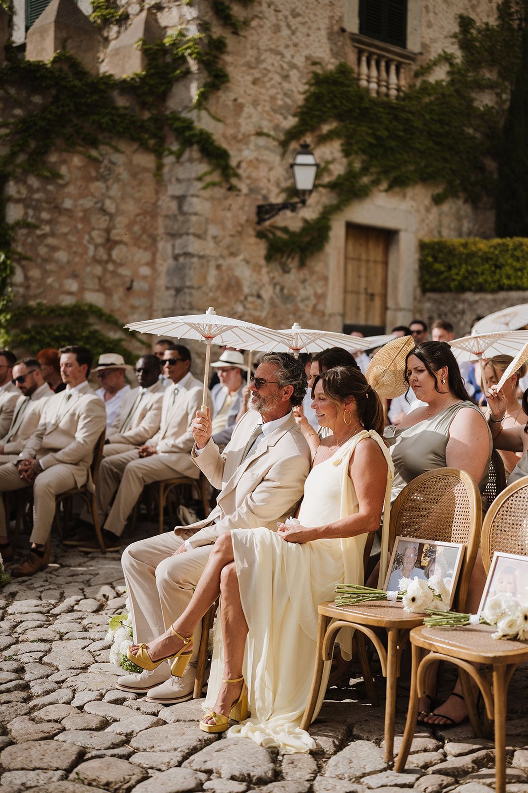 Wedding guests seated in rattan chairs at outdoor ceremony, woman in yellow dress beside man in beige suit, stone building behind.