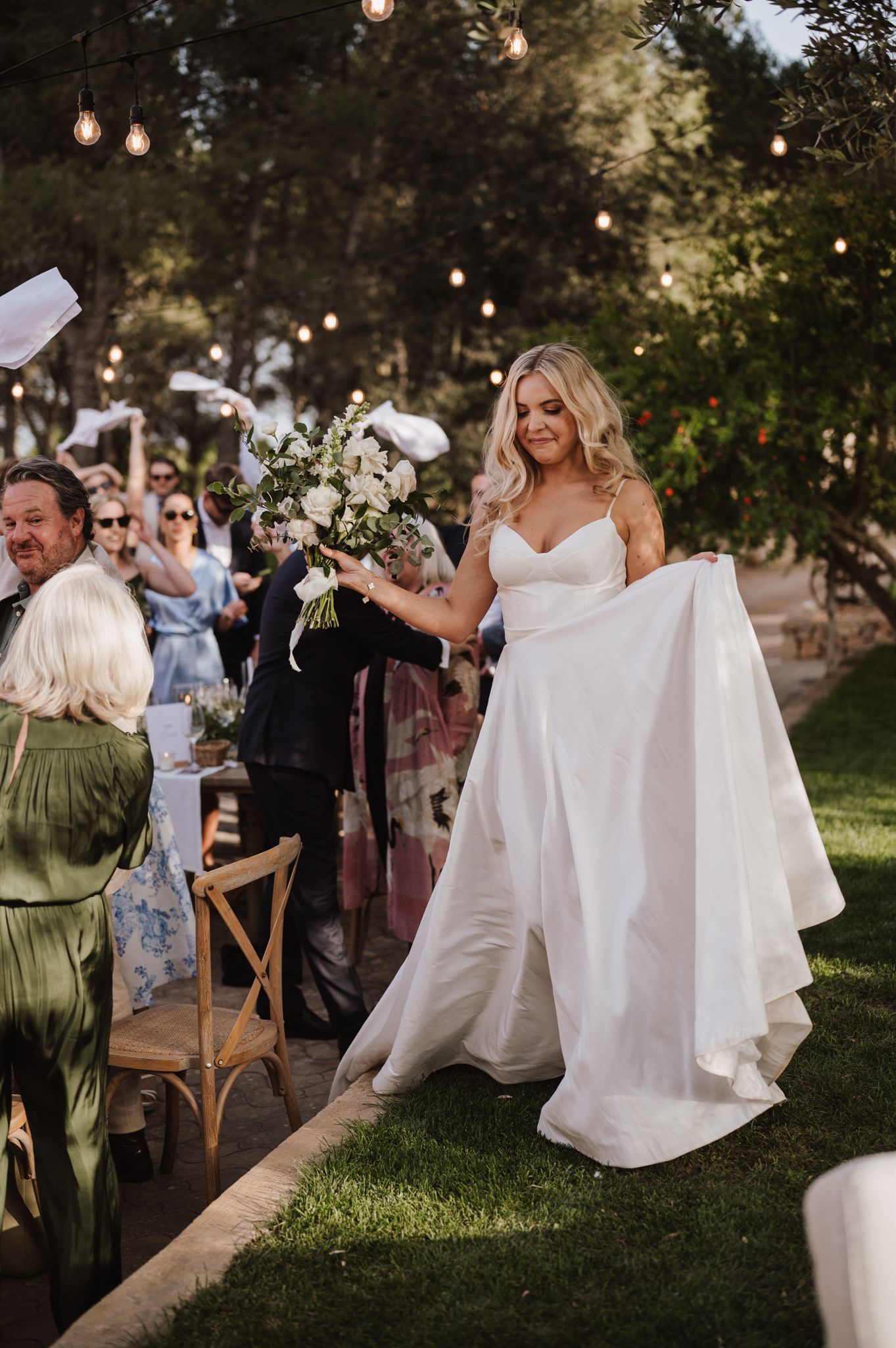 Smiling bride lifts her white gown while carrying bouquet under string lights at evening garden reception.
