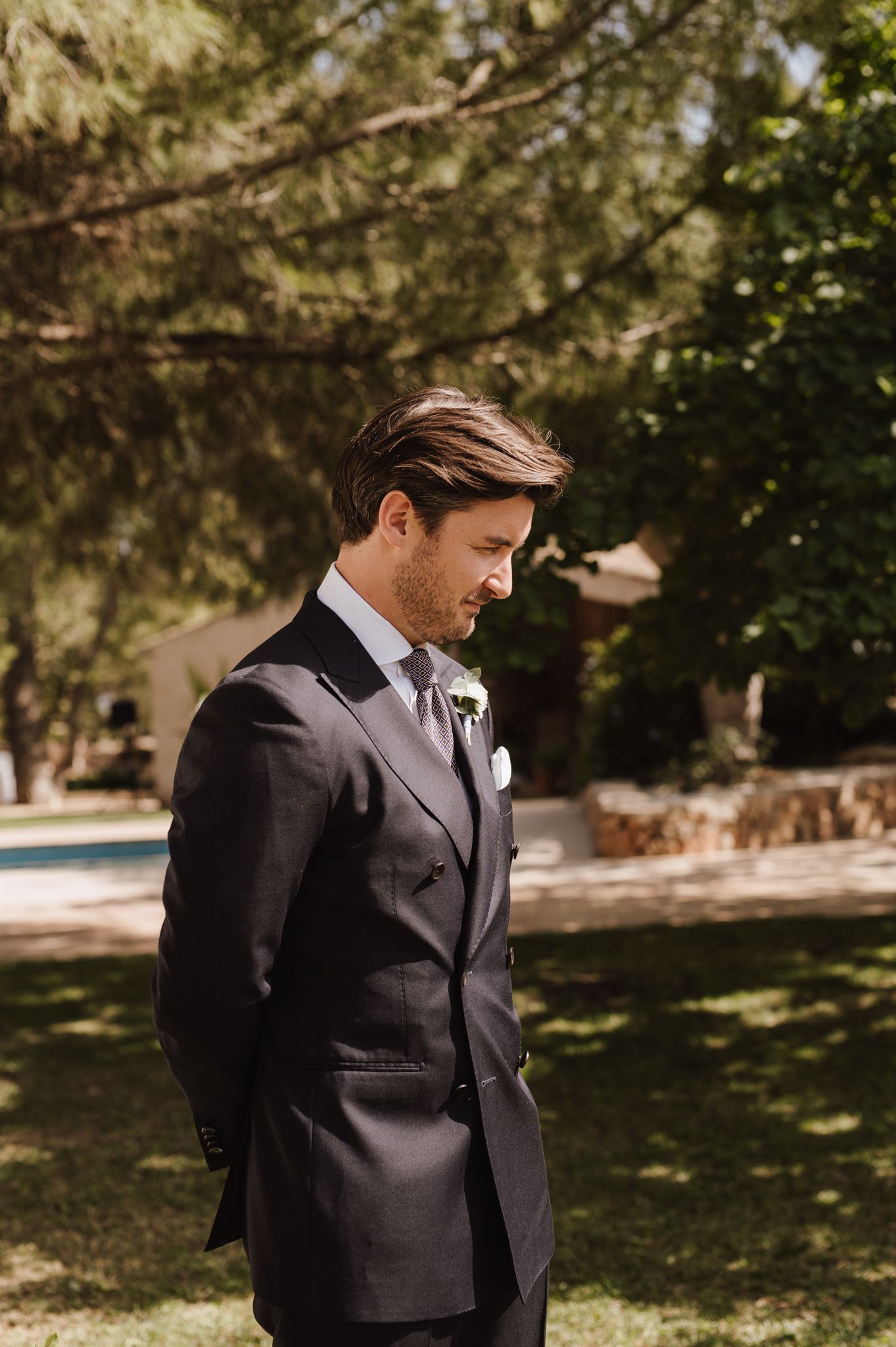 Elegant groom in a dark double-breasted suit with boutonniere standing outdoors in a garden.