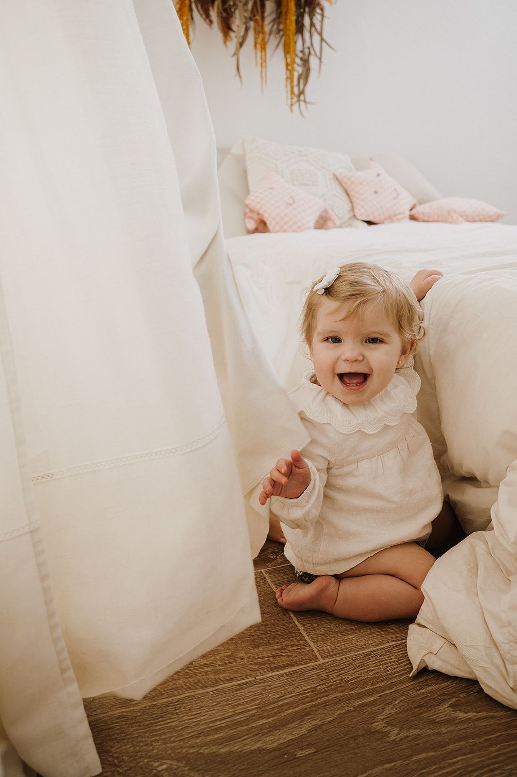 Happy toddler girl in white dress sitting on wood floor beside white bed with pink pillows, laughing joyfully.