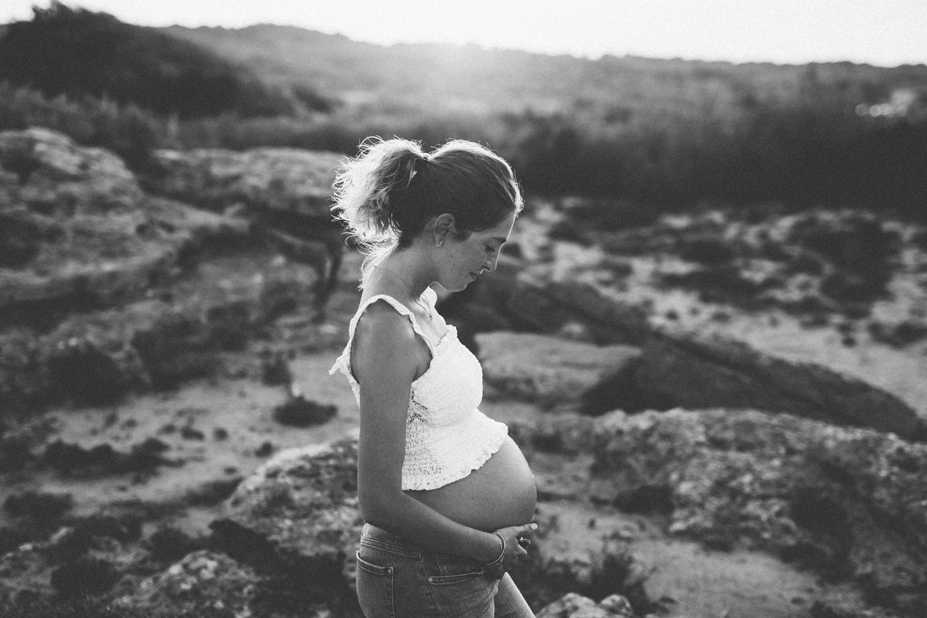 Black and white photo of a pregnant woman outdoors in a sandy landscape, cradling her baby bump in sunlight.